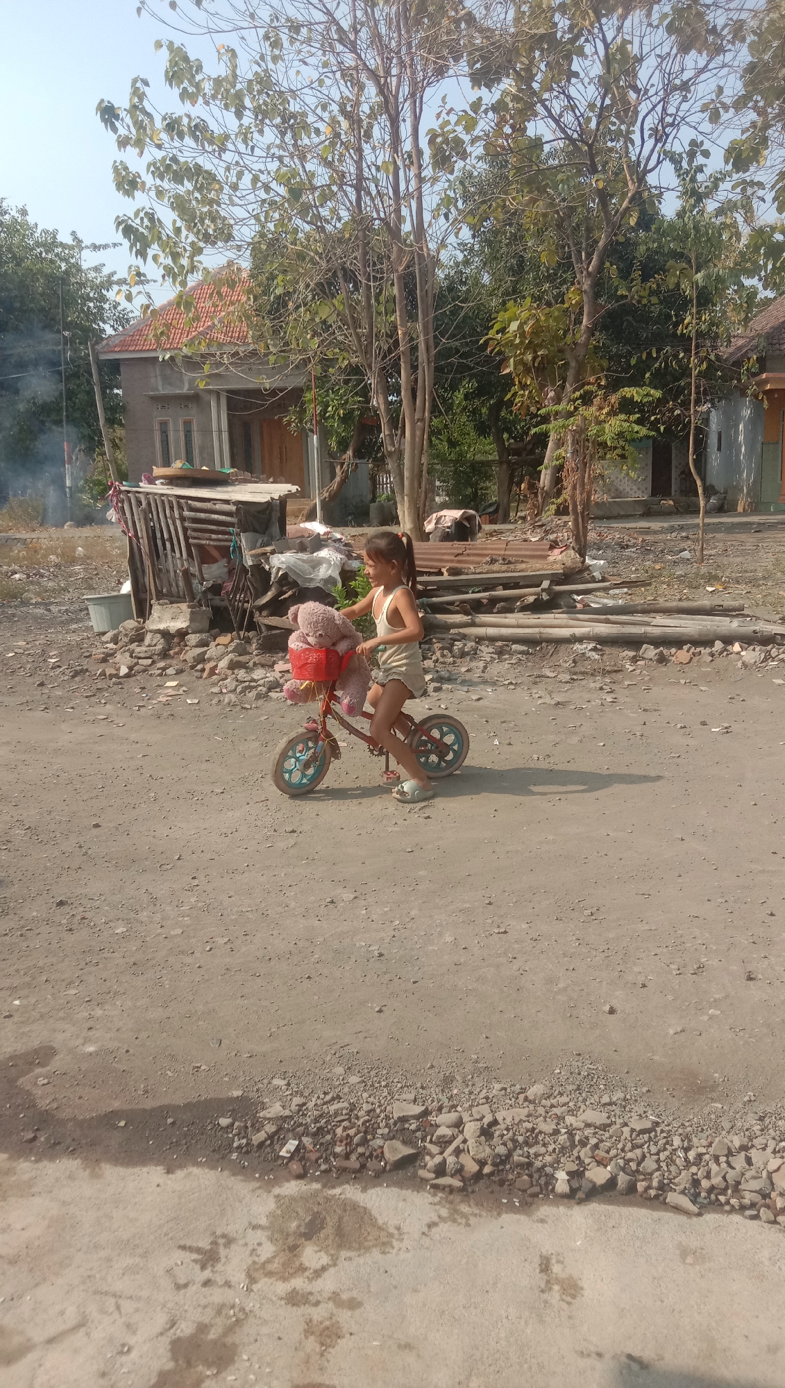 A young girl rides a red bicycle on a dirt road in what appears to be a rural or semi-urban area of Karang Anyar, Indonesia. The girl is wearing a white tank top and light-colored shorts, with her hair pulled back in a ponytail. She is smiling and looking to her left. Her bicycle has a red basket attached to the handlebars, which contains a large, pink teddy bear. The bicycle has blue rims on its wheels.
The environment is dry and dusty. In the background, there are several simple houses, some with red tile roofs. There are also trees, some of which appear to be bare or sparsely leafed, suggesting a dry season. Piles of wood and debris are scattered around, along with a large grey plastic bin. Smoke is visible in the far left background, possibly from a fire. The road is unpaved and appears to be made of dirt and gravel, with some patches of concrete or pavement at the bottom of the frame. The lighting indicates it is daytime, likely late morning or early afternoon, with strong shadows cast on the ground.