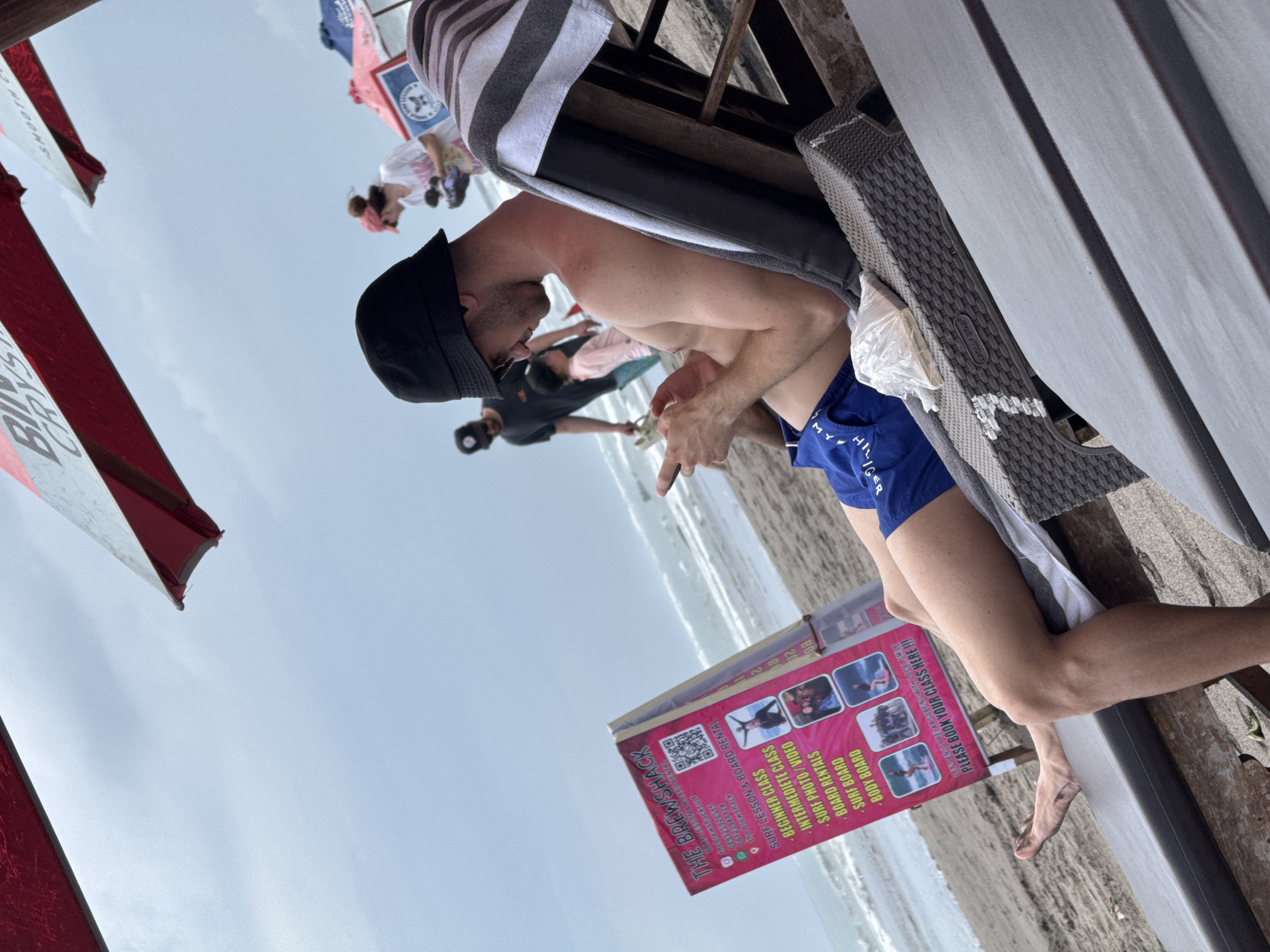 This image, taken in North Kuta, Indonesia, captures a casual daytime scene on a sandy beach under an overcast sky. The primary subject is a shirtless man wearing blue Tommy Hilfiger swim trunks and a black bucket hat, reclining on a grey and wicker beach lounger. He appears relaxed, looking down at an object in his hands. A striped towel is draped over the lounger.
In the background, other people are visible further down the beach near the ocean's edge. Partial views of red and white beach umbrellas or flags, possibly bearing text like "SMOOTH CO," "APOLLY STAR," and "BUD," frame the scene.
A prominent pink sign on the sand advertises "THE BREWSHACK," offering "SURF LESSON & BOARD RENTAL." The sign lists services such as "BEGINNER CLASS," "INTERMEDIETE CLASS," "SURF PHOTO/VIDEO," and "BOARD RENTALS" for "SURF BOARD" and "BODY BOARD." It also displays contact information including phone numbers (089525920515, 08123947124) and an Instagram handle (@thebrewshacksurf). The motto "LET'S ENJOY RELAX SURFING WITH LOCAL SMILE" and the call to action "PLEASE BOOK YOUR CLASS HERE !!!" are also visible. The setting suggests a leisurely beach environment focused on relaxation and water activities.