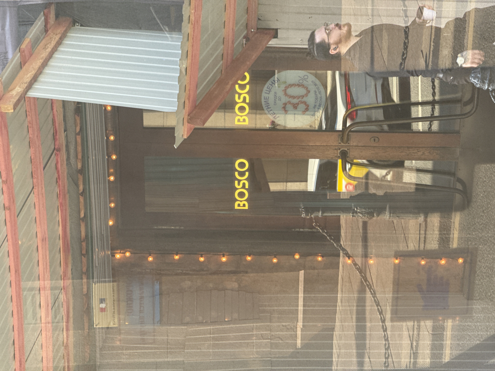 The image depicts a street-level view of a commercial establishment in Saint Petersburg, Russia, as indicated by a sign with the French flag and Cyrillic text. The establishment's entrance features a wooden door with large glass panes, through which the interior and some of the street are visible due to reflections. Yellow lights are strung along the door frame, illuminating the entrance. The name "BOSCO" is displayed prominently in large yellow lettering on the glass doors.
To the right of the entrance, a circular sign with Cyrillic text and the number "30%" is affixed to the glass, suggesting a sale or discount. The text above "30%" appears to read "ЛЕТНИЕ ЦЕНЫ" (Summer Prices) and below it, "НА ТОВАРЫ" (On goods). The bottom text reads "ВЕСНА-ЛЕТО 2025" (Spring-Summer 2025).
A man wearing glasses and a beard, with his hair tied back in a bun, is walking past the establishment from right to left. He is dressed in dark clothing and is holding a white cup in his right hand. He appears to be the only person in the immediate vicinity.
Reflections in the glass show a building facade with vertical lines and what appears to be a metal roof with reddish-brown structural elements. A chain barrier is visible across the sidewalk in front of the entrance. In the background, a white car with red taillights is partially visible, suggesting a street or parking area. The overall lighting suggests it is daytime, with sunlight creating reflections and highlights.