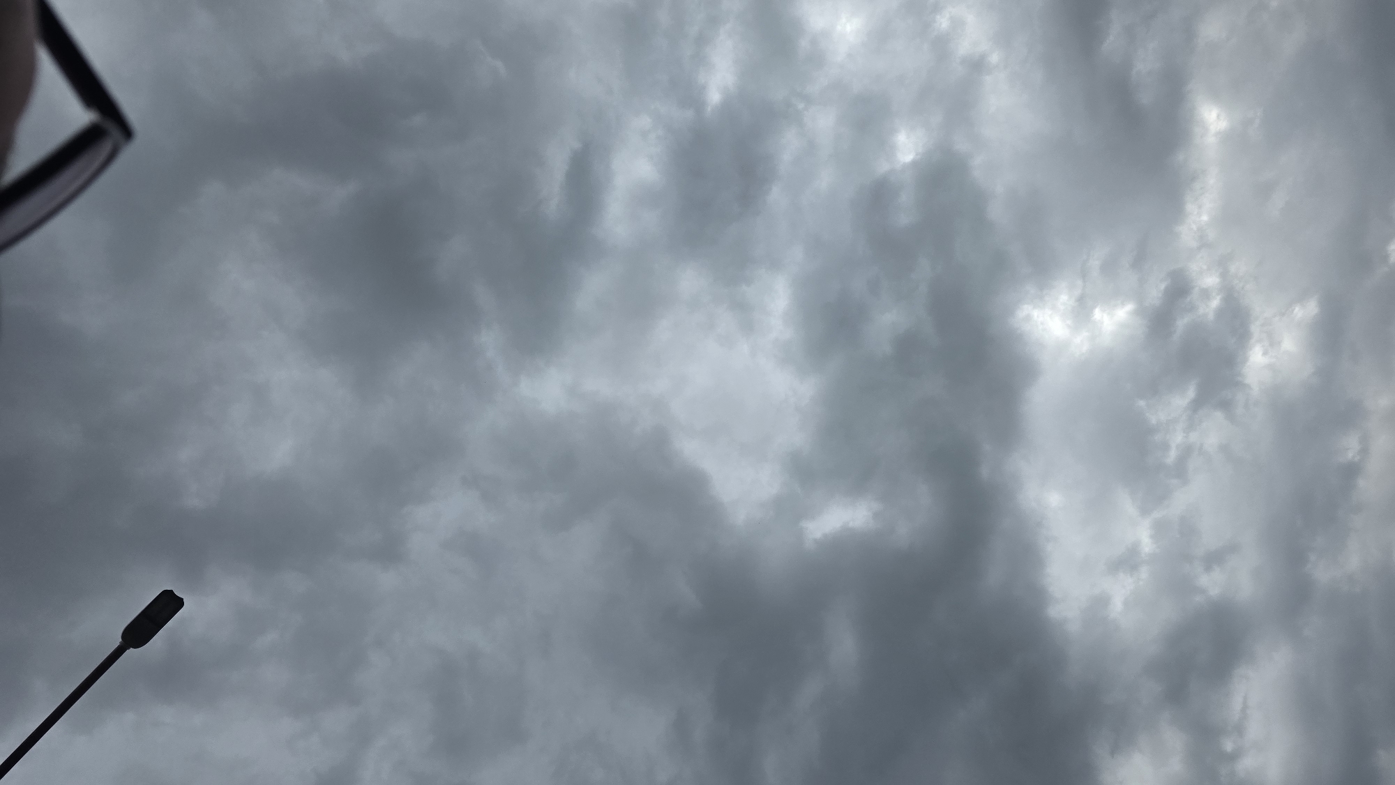 A low-angle shot of a cloudy sky, possibly taken in Blagoevgrad, Bulgaria. A lamppost is visible in the lower left corner of the frame. A pair of eyeglasses is partially visible in the upper left corner. The sky is overcast with grey clouds.