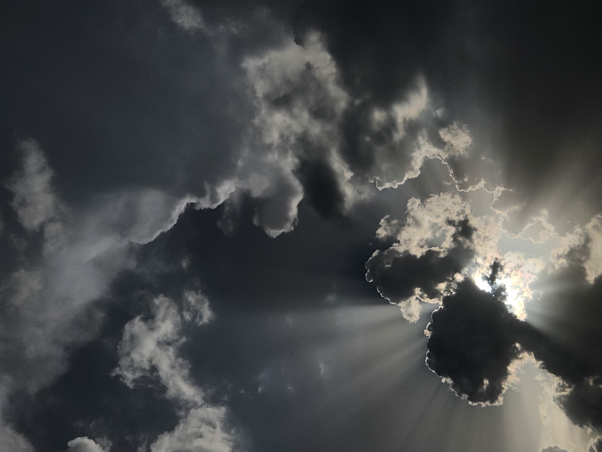 The image displays a dramatic sky filled with dark, turbulent clouds. Sunbeams pierce through gaps in the clouds, creating a crepuscular ray effect. One prominent dark cloud formation in the lower center appears to be lit from behind by the sun, with rays emanating outwards. The overall mood is somber and powerful. The location is identified as Altamonte Springs, United States of America. There are no people, subjects, objects, or actions depicted in this image, only natural atmospheric phenomena.