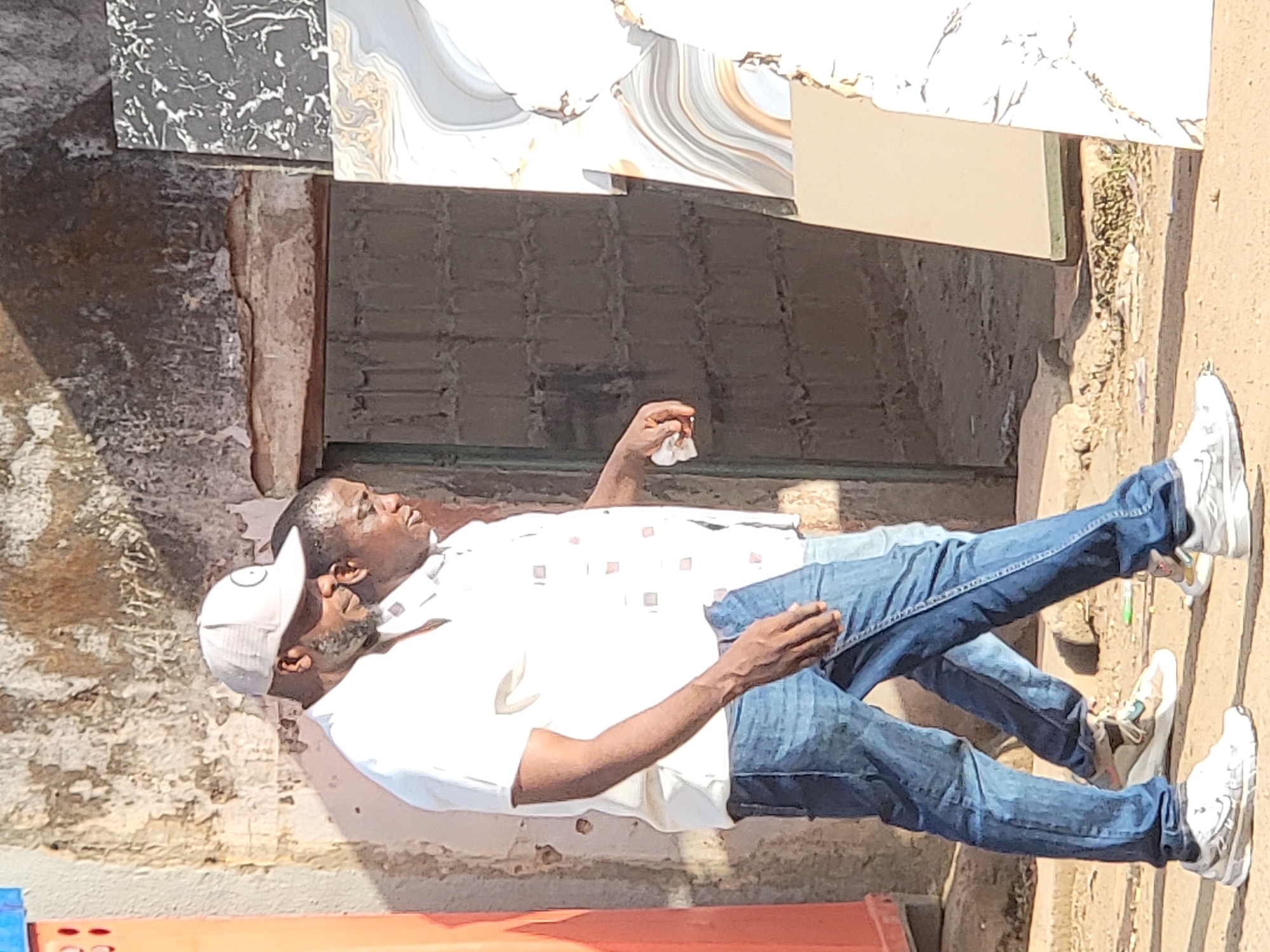 A man wearing a white shirt, blue jeans, and white sneakers stands on a dirt sidewalk in Maiburiji, Nigeria. He also wears a grey baseball cap. He holds a white cloth in his right hand. Behind him, a stack of tiles rests against a building with peeling paint and a closed door.