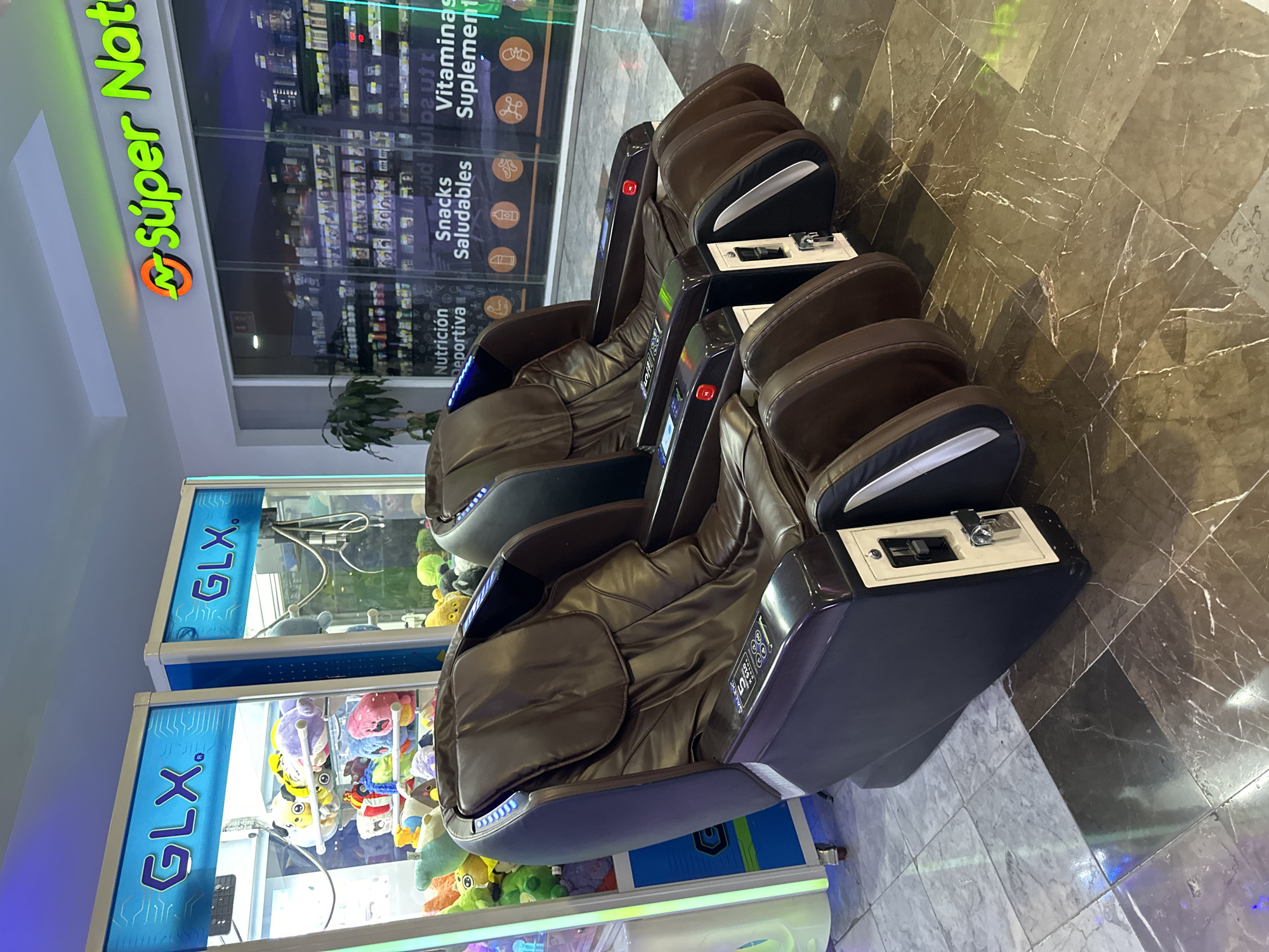 The image depicts a brightly lit indoor setting, likely a shopping mall in Huixquilucan de Degollado, Mexico. Three identical massage chairs are arranged in a row, angled toward the right. The chairs are brown leather with black and white accents and coin-operated mechanisms. To the left, two claw crane machines are visible, labeled "GLX" in large, vertically oriented blue letters. The cranes contain stuffed animals. In the background, a storefront with the sign "Súper Nat" and windows displaying health products (vitamins, snacks) are evident. The floor is tiled with a dark, marble-like pattern.