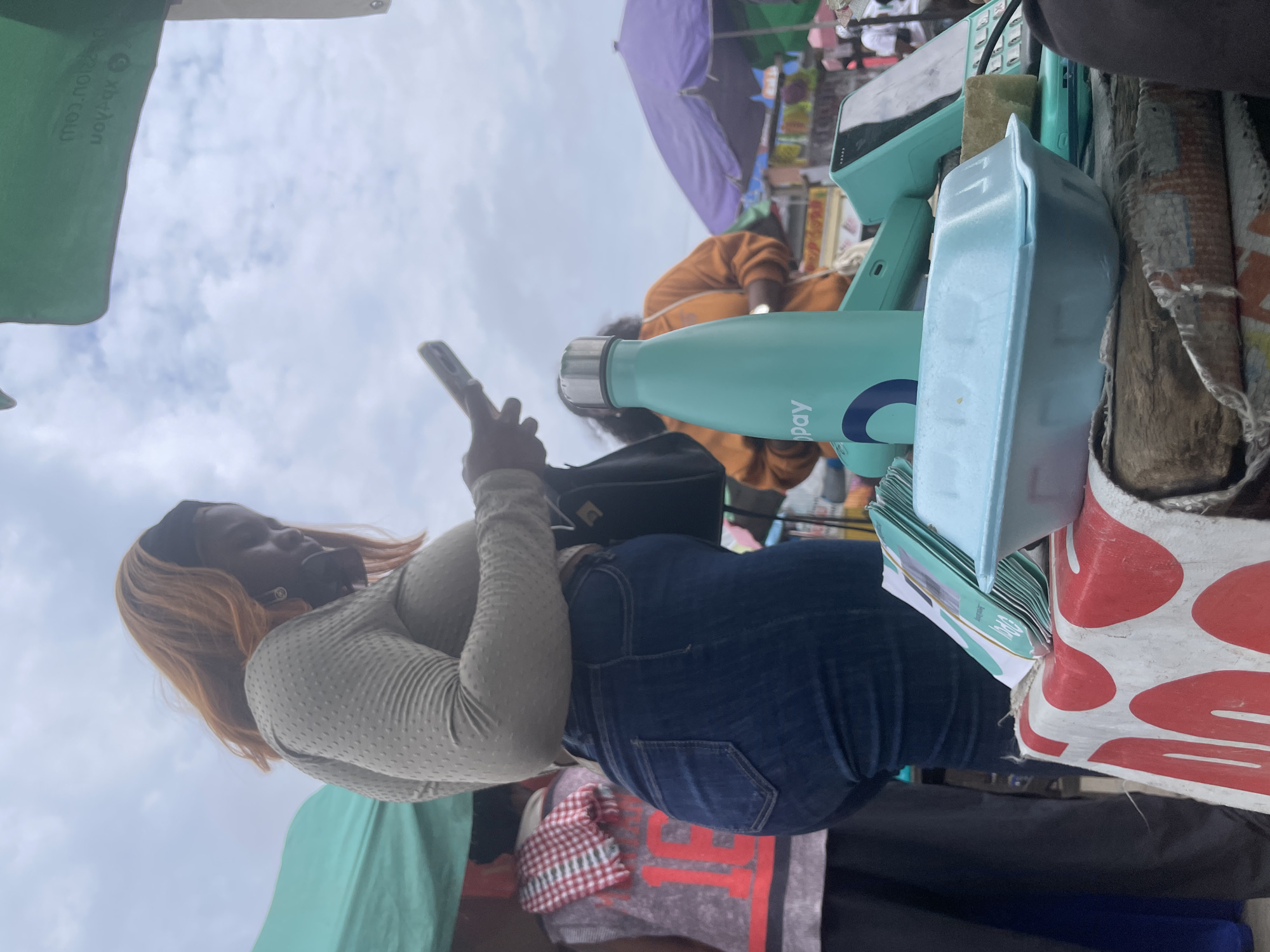 In Ajah, Nigeria, a woman with auburn hair stands outdoors, looking at her phone. She is wearing a light grey, long-sleeved top with a textured pattern and dark blue jeans. To her left, another person is partially visible, wearing a grey shirt with red lettering and a checkered fabric draped over their shoulder. In the foreground, a table is set up, covered with a white cloth featuring red circular designs. On the table, a light blue styrofoam container, a stack of pamphlets or brochures, and a teal-colored water bottle with a silver lid are visible. The water bottle has a logo that includes the word "CoPay." In the background, other individuals, market stalls, and umbrellas can be seen, suggesting a bustling outdoor market environment. The sky is cloudy.
