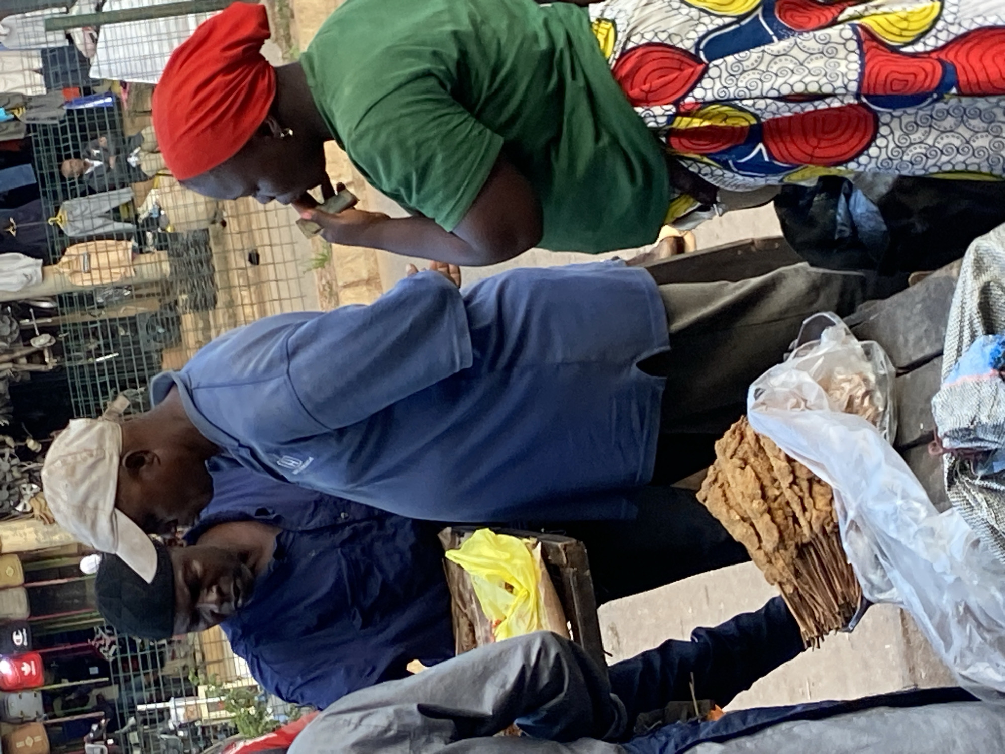 In Bukuru, Nigeria, at a street market, two men and a woman are gathered around a wooden stand. The man on the left, wearing a black skullcap and a navy blue shirt, looks down. To his right, a man in a blue t-shirt and a light-colored baseball cap gestures with his left hand. He has a prominent stomach. The woman on the far right, dressed in a green t-shirt and a vibrantly patterned skirt, has a red headwrap and is holding a small object to her mouth. In front of them, on the wooden stand, are skewers of fried food, partially covered by a clear plastic bag, and a yellow plastic bag. Behind them, a metal fence displays various clothing items for sale. The ground is paved.