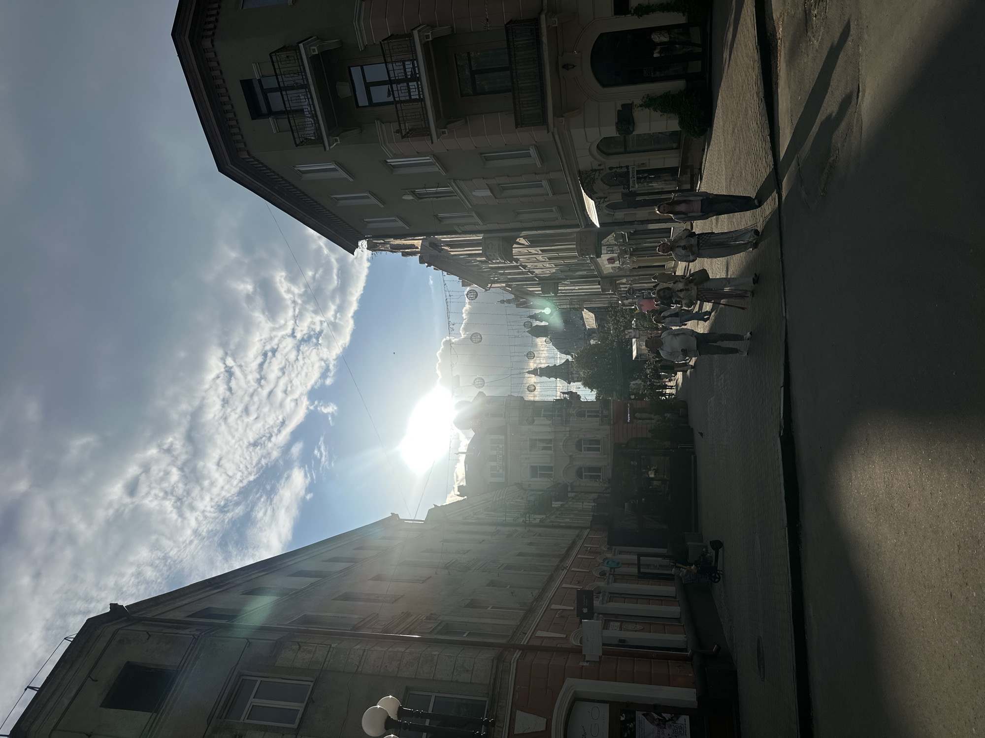 The image captures a street scene in Ternopil, Ukraine, under a bright sky with the sun obscured by clouds. Tall buildings line both sides of the street, typical of European urban architecture, with details like balconies and ornamented facades. A group of people walks along the sidewalk on the right side of the frame. Overhead, decorative light fixtures, possibly globe-shaped, are strung across the street. The street itself appears paved and wet in areas, possibly from recent cleaning or weather. Shadows are cast on the street due to the sunlight.