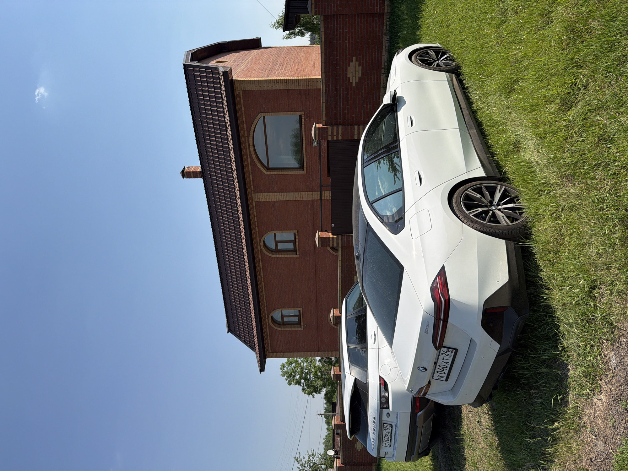 The image depicts two white vehicles, a house, and a fence under a clear blue sky on a bright, sunny day.
In the foreground, a white BMW 220i sedan (license plate K 040 XT 24 RUS) is parked on a grassy area, angled slightly towards the right. Its rear and right side are visible. The car features black multi-spoke wheels and a black lower rear bumper fascia. Behind and to the left of the BMW, a white Range Rover Evoque SUV (license plate C 700 BX 124) is partially visible, parked on the grass.
In the mid-ground, a two-story house with red brick walls and a dark brown tiled roof is prominent. The house features a brick chimney on the roof and multiple arched windows, including two smaller ones on the left side of the visible facade and one larger central window on the right. A brick fence, matching the house's brickwork, extends across the property front, with a dark brown gate visible.
The ground where the vehicles are parked is covered in green grass, appearing somewhat unkempt. The sky above is clear and light blue, with a single small white cloud visible in the upper right quadrant. Green foliage and trees are visible in the background on both the left and right sides of the property, and power lines with poles are discernible in the far left background.
Based on the Russian license plates "24 RUS" and "124 RUS" which correspond to Krasnoyarsk Krai, the location of this scene is within the city of Krasnoyarsk, Russia, or its immediate vicinity.