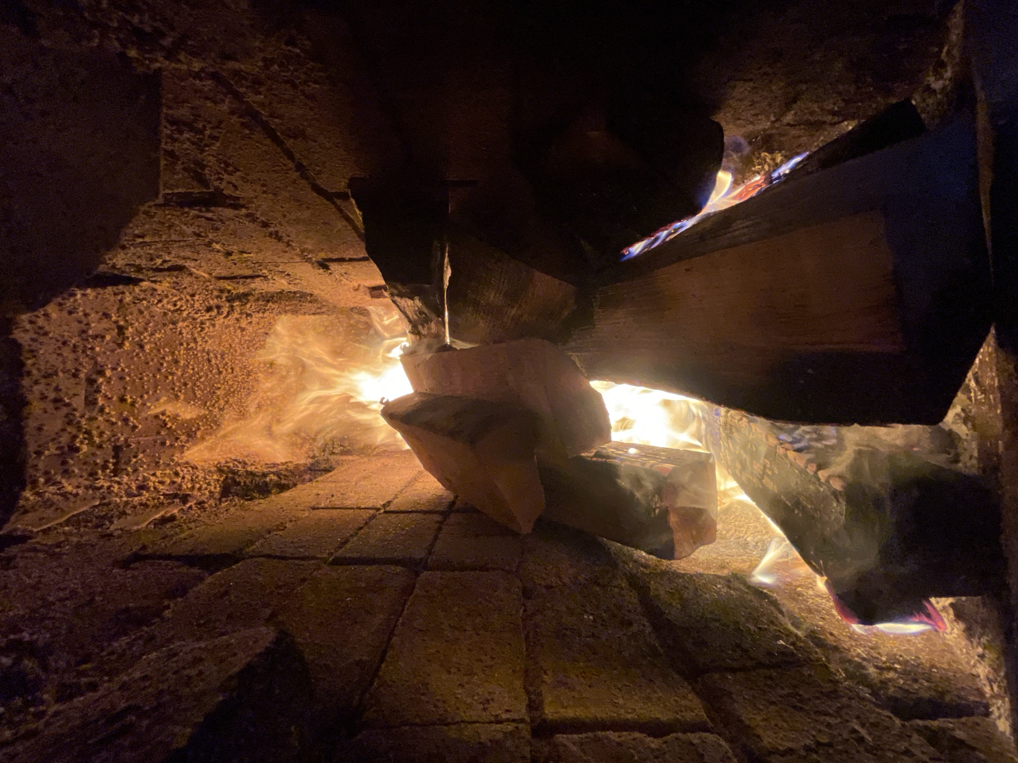 The image shows a fire burning inside a brick oven. The oven walls appear rough and porous, and the floor is made of rectangular bricks. Flames are visible between and around stacked pieces of wood. The location in the city of Sestroretsk, Russia is not determinable.