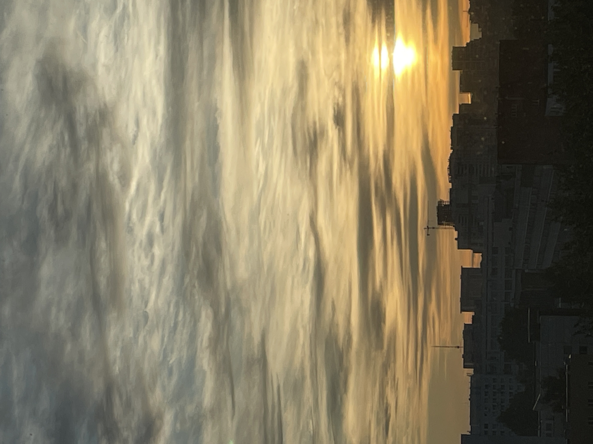 The image shows a landscape with the city of Izhevsk, Russia silhouetted against a golden, light-filled sky that fades into lighter, whiter clouds. The buildings are dark, and a construction crane is visible. The lighting is such that it appears to be sunrise or sunset.