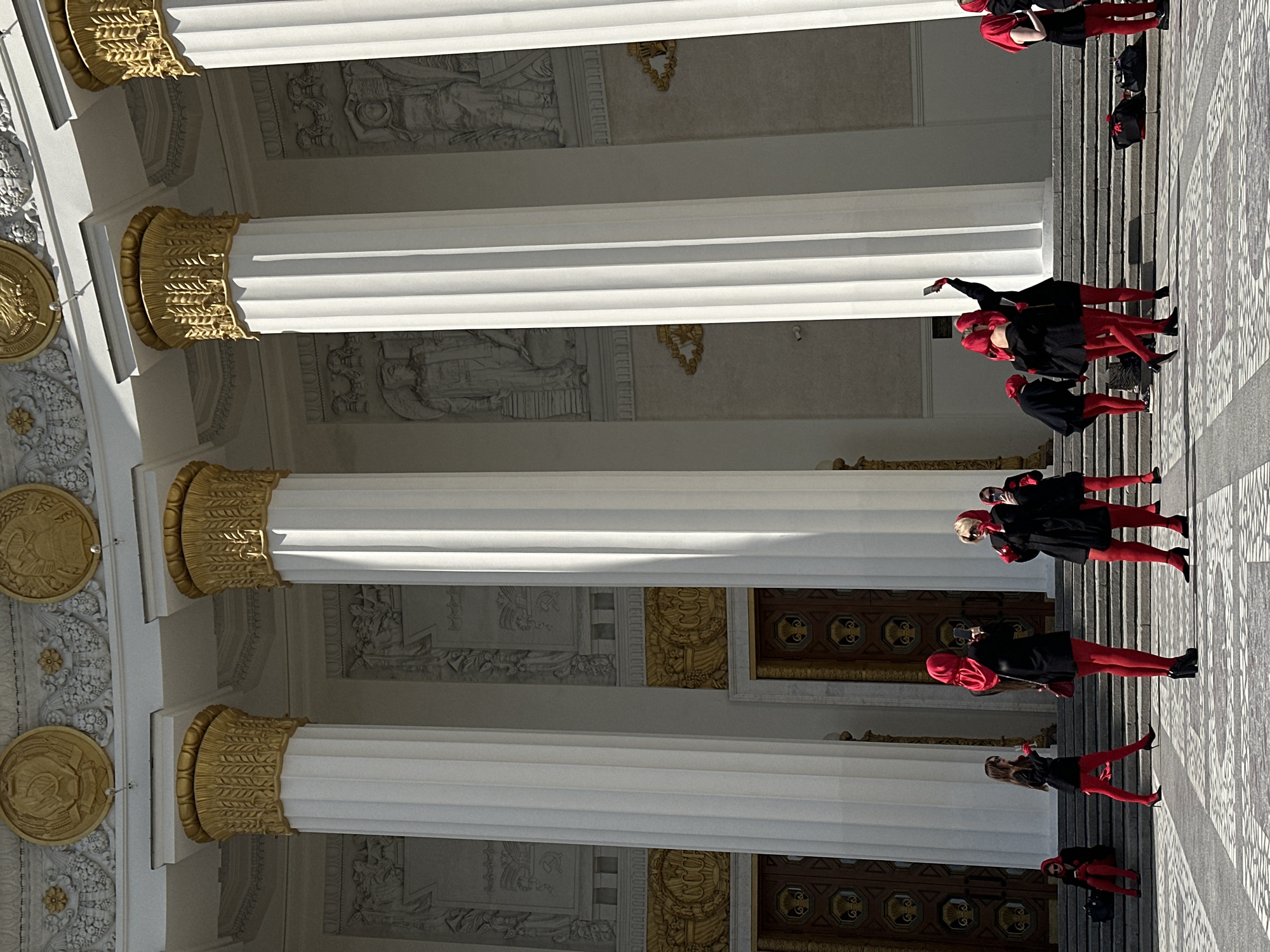 The image captures a group of individuals dressed in black coats and striking red tights, posing on the steps of what appears to be an architectural building. The location is Unknown, Unknown. The building is adorned with large, white columns capped with ornate gold detailing. These columns line a decorative facade. The steps the group stands on lead to a paved area. The individuals appear to be interacting, with some holding phones and possibly taking pictures.