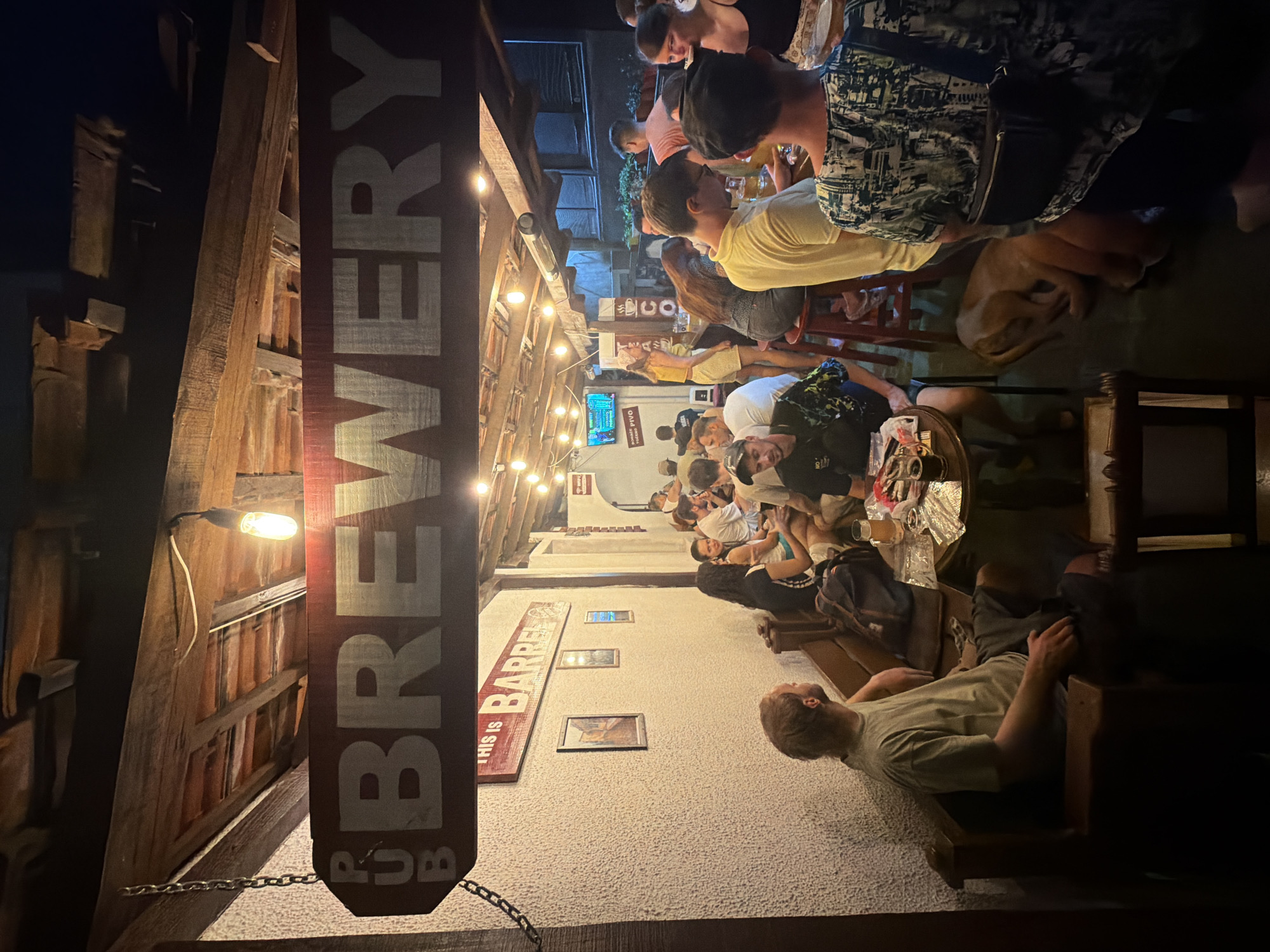 This is a medium shot of the outdoor seating area of a brewery or pub in Bar, Montenegro, at night. A wooden sign above the entrance reads "PUB BREWERY" in white lettering. Below this, another sign reads "THIS IS BARREL". The area is lit by string lights and an overhead bulb, creating a warm, ambient glow.
Several people are seated at tables and at the bar, enjoying drinks and conversation. In the foreground, a man with blond hair wearing a tan t-shirt and dark shorts sits on a wooden bench, looking away from the camera. A dog is curled up on the floor near a table in the middle ground. Further back, a group of people are standing and sitting around a bar area, where drinks and food are visible on the counter. The walls are textured and light-colored, and the ceiling is made of exposed wooden beams. The overall atmosphere appears casual and relaxed.