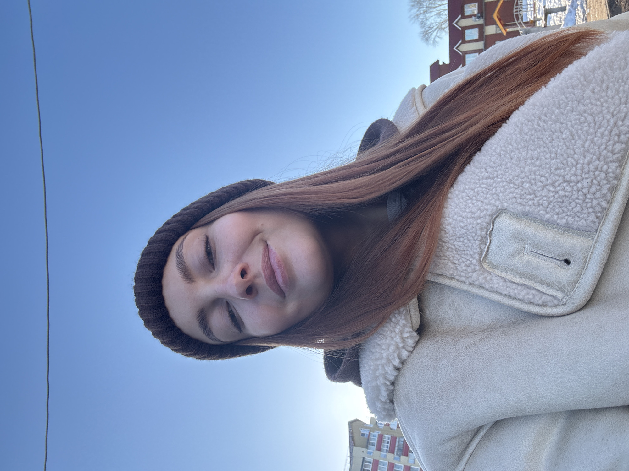 A young woman with auburn hair and wearing a brown beanie and a light-colored jacket is seen from a low angle, seemingly taking a selfie on a bright, sunny day. Her eyes are closed in a slight smile. Behind her, a clear blue sky dominates the upper portion of the frame. Buildings are visible in the background, suggesting an urban environment. The architecture and color scheme suggest it is the city of Chaikovsky, Russia. A thin, dark wire is seen against the sky, likely a power or communication line.