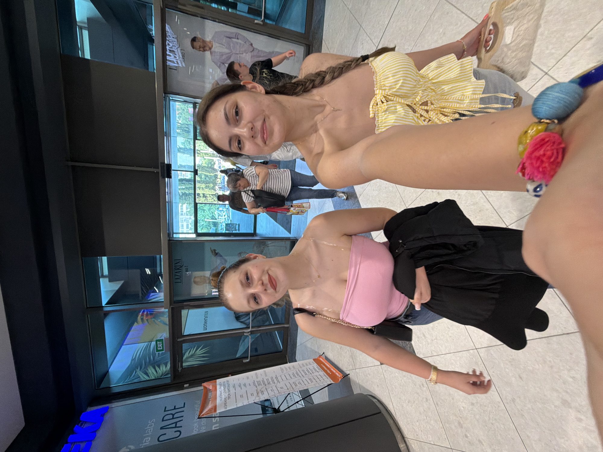 The image is a selfie featuring two young women in what appears to be a modern indoor public space, likely a shopping mall or similar commercial building, in Burgas, Bulgaria. The woman on the left (viewer's left) wears a pink strapless top, jeans, and has a black jacket draped over her arm. Her light brown hair is styled in an updo, and she smiles subtly towards the camera. The woman on the right, closer to the camera, sports a yellow and white striped strapless top with decorative ties, light-colored trousers, and a long braid. She also wears a necklace and a vibrant beaded bracelet on her wrist. Both subjects appear relaxed and content.
The background shows a light-tiled floor and large glass walls, through which other people and reflections of an outdoor environment with greenery and buildings are visible, suggesting it is daytime. A freestanding banner in the mid-ground displays a long string of Cyrillic text, including "HAUCHANDSVIREA КУЗИКАЛНО И СЦЕНИЧОВОО ТРОФ ПАРОВНИЛСЯ". Other visible text includes "Ha labs CARE" and "look beautiful" on a wall sign, an "EXIT" sign, and "LAZARINI" and "ШОПИНГЪТ Е В" (Shopping is in/on) on reflective surfaces, reinforcing the commercial setting.