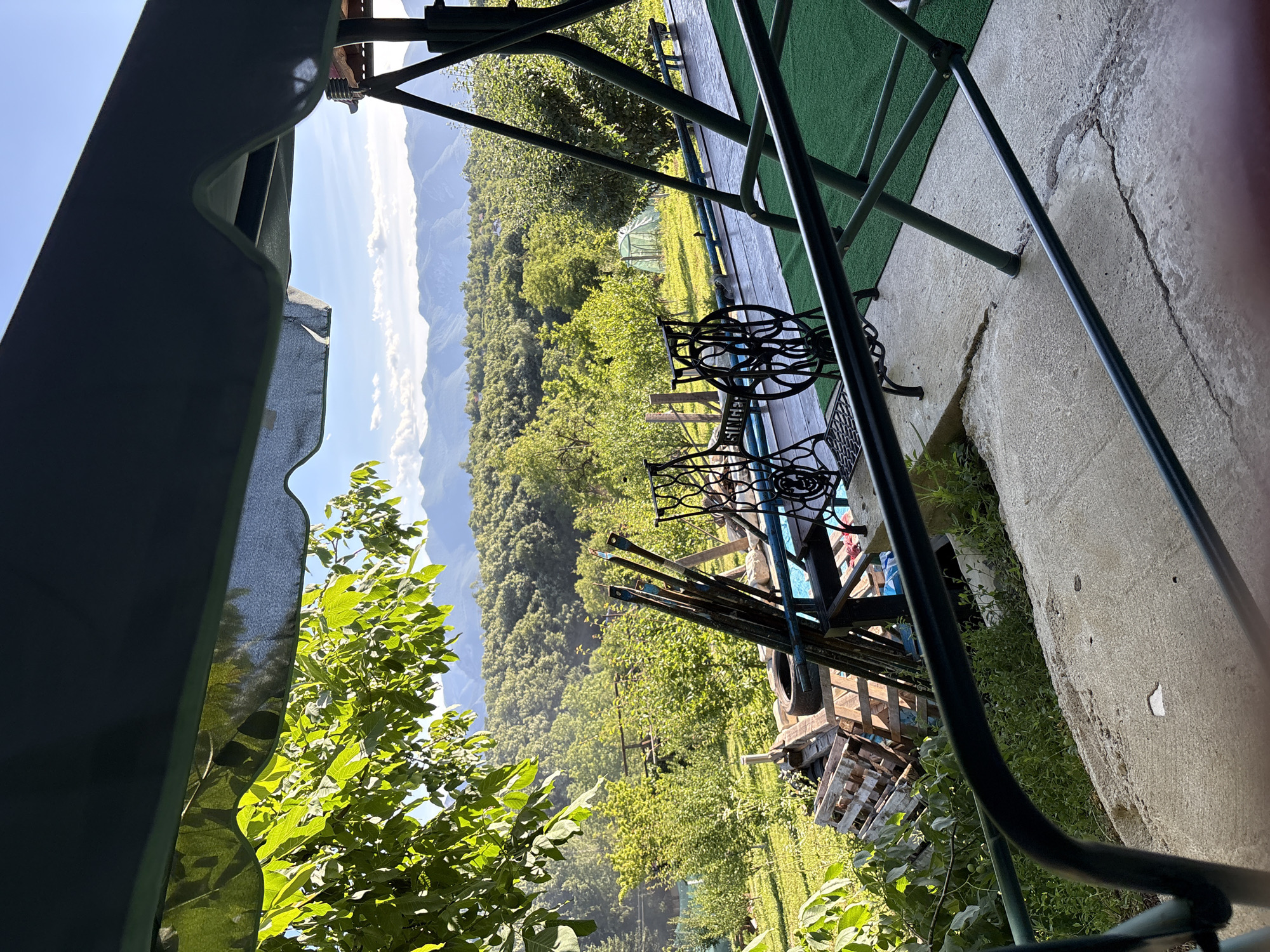 The image depicts a view from a covered patio or balcony overlooking a rural landscape. In the foreground, a green metal swing set frame is visible, with parts of its legs and structure extending into the frame. In front of the swing set, there are two decorative wrought-iron bases of old sewing machines. To the left, a concrete patio is visible with some patchy grass and weeds growing through cracks. Further into the scene, stacks of wooden pallets, a tire, and some long metal poles are piled up. Beyond this area, a lush green lawn extends towards a wooded hillside. In the middle ground, there is a small green greenhouse. The background is dominated by a vast expanse of dense green forest that stretches towards rolling hills and distant mountains, under a clear blue sky with scattered white clouds. The overall impression is of a peaceful, natural setting, likely in a rural area outside of a city. Without further information, it is not possible to determine the specific location within Vratsa, Bulgaria. There are no people or animals visible in the image.