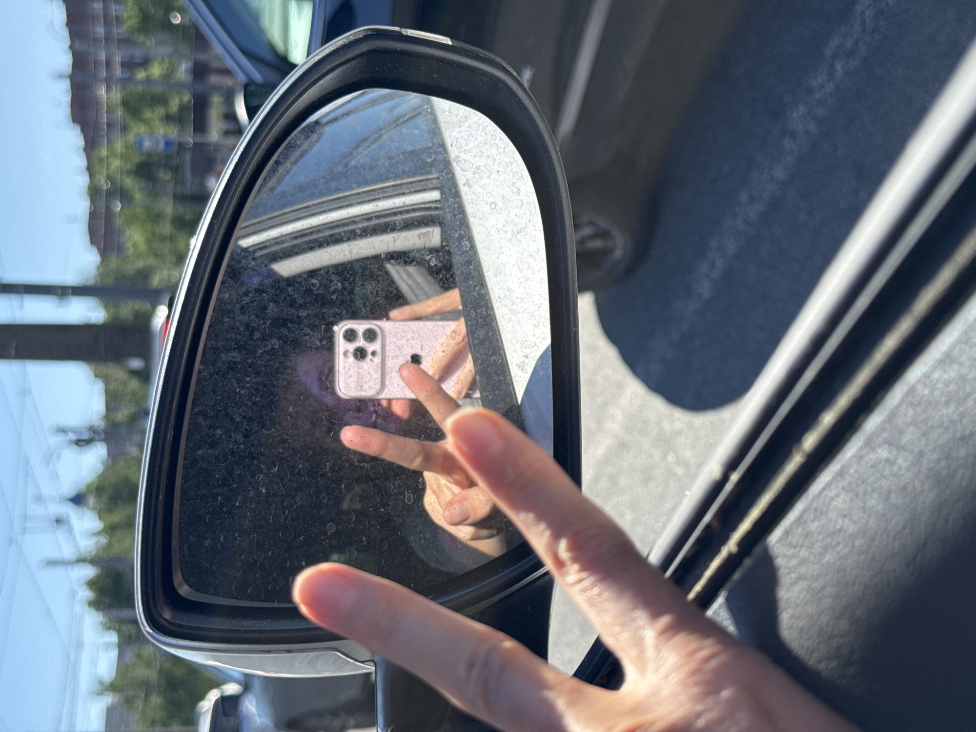 The image presents a first-person view from a vehicle in Yukki, Russia, on a bright, sunny day. In the immediate foreground, a light-skinned hand extends from the lower left, forming a peace sign gesture. The central subject is a black-framed side-view mirror, heavily obscured by water spots or dust, reflecting the photographer. The reflection shows a person holding a pink iPhone, identifiable by its triple-camera array, with their reflected hand also forming a peace sign, obscuring their face. The background, visible through and around the mirror, reveals a blurred urban or suburban environment under a clear blue sky. Distant structures include a prominent dome or spire, various other buildings, green foliage, utility poles with overhead lines, and parts of other vehicles on an asphalt road. Strong shadows are cast, indicative of direct sunlight.