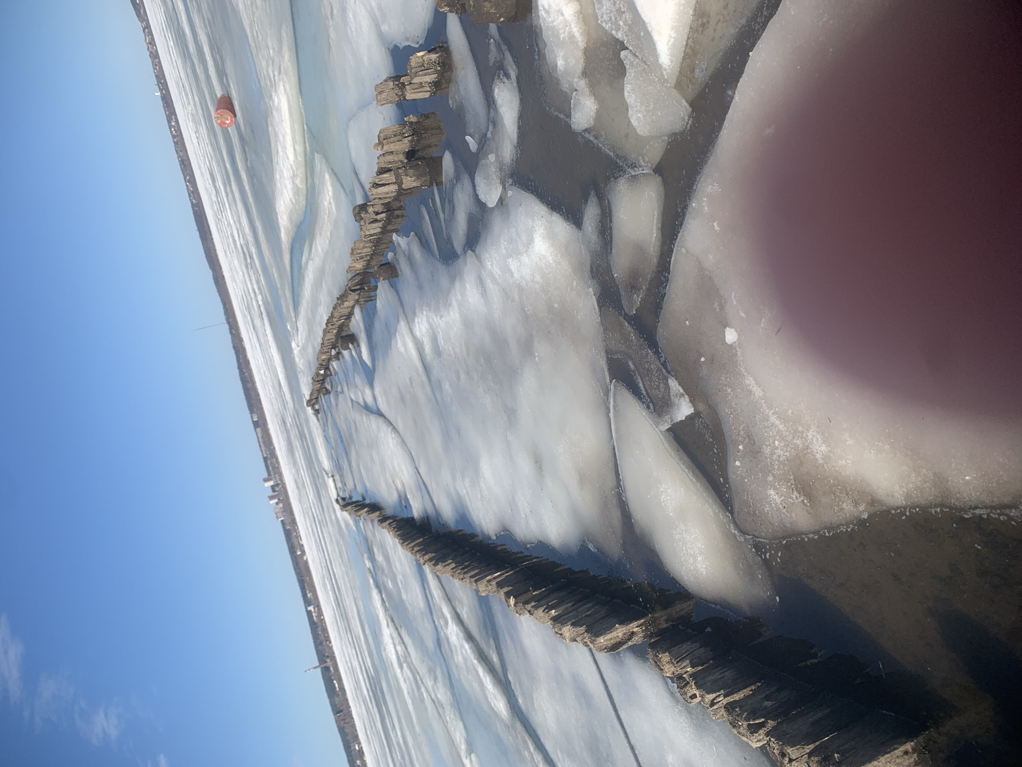 The image depicts a frozen body of water, likely a river or lake, with ice formations and wooden structures extending across the water's surface. The location is suggested to be in or near Izhevsk, Russia. The scene includes a partially frozen body of water, wooden posts or breakwaters extending into the water, and a horizon line indicating a shoreline with potential buildings or structures in the distance. The sky is blue, suggesting a sunny day. There is a reddish-orange object that could be a buoy or marker floating in the distance. The presence of ice and the wooden structures suggest the possibility of human intervention or modification of the environment.