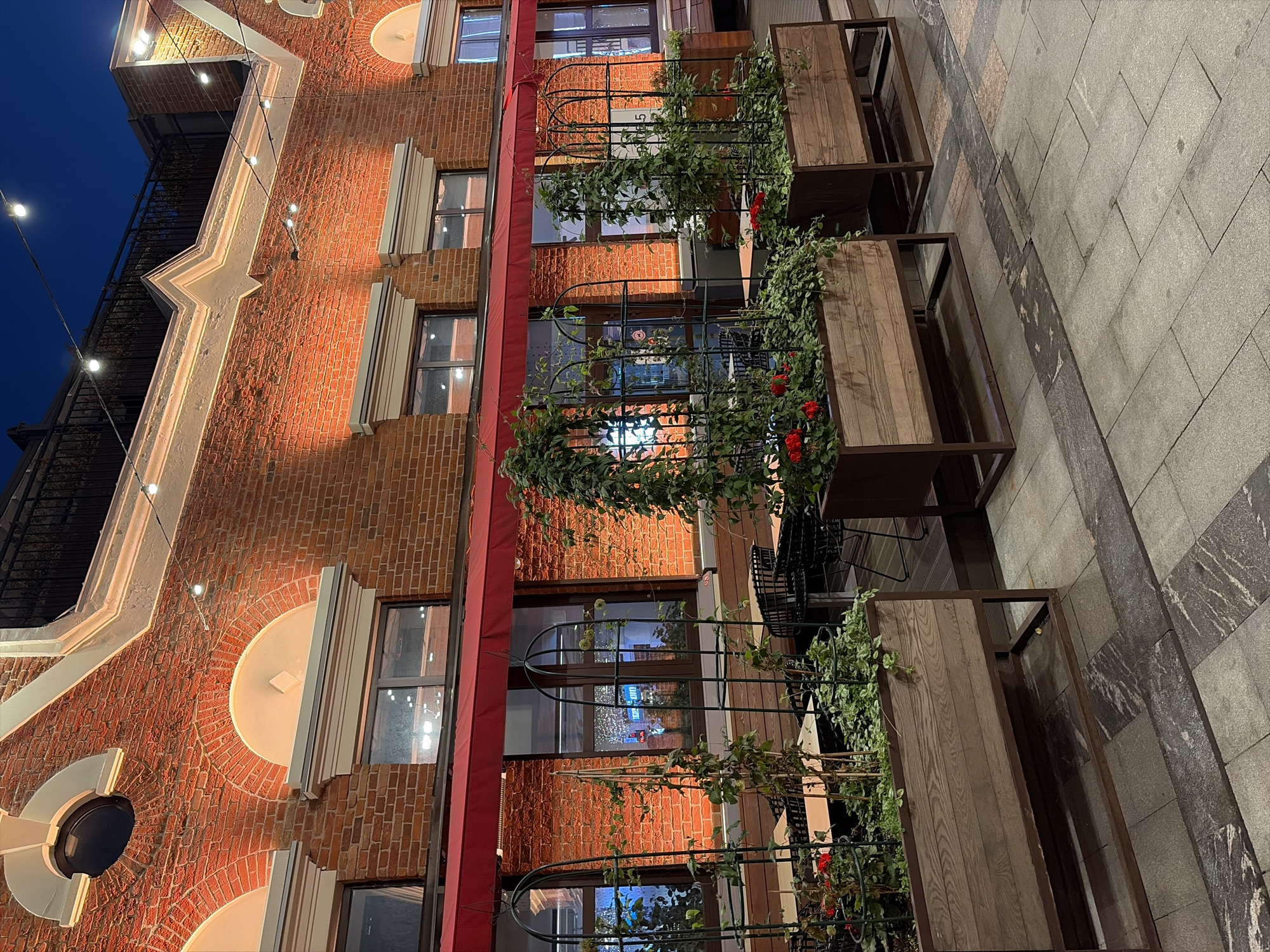 The image depicts the exterior of a brick building at night, illuminated by warm orange lights. The building features arched windows and a red awning extending across the facade. In front of the building, several large wooden planters with metal trellises are filled with greenery and red flowers. Outdoor seating areas with tables and chairs are visible behind the planters. The ground is paved with stone tiles. String lights are visible overhead, and the sky is dark. This scene is located in Moscow, Russia, specifically at the Central Market (Tsentralnyy Rynok) on Rozhdestvensky Boulevard, a popular food market and social space.