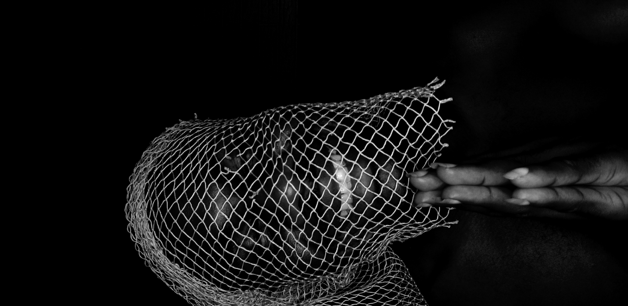 This black and white photograph presents a stark, close-up composition of a person partially obscured by a mesh net. The central focus is the left side of a person's head, which is covered by a coarse, white, diamond-patterned net, similar to those used for packaging.
Through the netting, dark skin is visible. The person's mouth is open, revealing prominent white teeth, which could suggest an expression of discomfort, surprise, or even a suppressed smile. The rest of the face, including the eyes, is largely concealed by the net's pattern and shadows, adding to a sense of anonymity or entrapment.
To the right of the covered face, a person's hand with neatly manicured, light-colored nails is positioned horizontally. The fingers are held close together, and the hand appears to be resting on a reflective surface, creating a subtle mirrored reflection below it. The hand's interaction with the net is ambiguous; it could be holding it, pushing against it, or simply resting nearby.
The background is entirely black, providing no environmental context and emphasizing the subjects. The high contrast of the black background against the white net and the illuminated teeth makes the image visually striking. The black and white palette enhances the dramatic and symbolic nature of the scene, evoking themes of restriction, concealment, or inner struggle. No text is visible in the image.