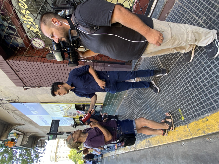 The scene captures a street interview in an urban environment, specifically identified as Buenos Aires, Argentina, due to the presence of "PAGO FACIL" and "Western Union" signage. Three individuals occupy the foreground on a sidewalk composed of metal grates with a yellow painted curb line.
On the left, an older light-skinned woman with light hair pulled back and wearing glasses, a purple t-shirt, denim shorts, and black sandals, is actively speaking, with her mouth open and hands raised in a gesture. She carries a black crossbody bag.
In the center, a light-skinned male reporter, mid-20s to early 30s, dressed in a dark blue button-down shirt, dark blue jeans, and black athletic shoes, holds a red microphone branded "GEN" towards the woman. He also holds a dark mobile device in his left hand and wears a black earpiece in his right ear, facing the woman.
On the right, a light-skinned male cameraman, mid-30s to 40s, with a beard, is operating a professional video camera positioned on his right shoulder. He wears a black t-shirt with a white stripe, tan cargo pants, black athletic shoes, white wired earbuds, and black safety goggles around his neck. He faces slightly past the reporter.
The background features multiple building facades. An older structure with an ornate balcony is visible on the far left. Adjacent to it, a building wall displays large advertisements for "PAGO FACIL" and "Western Union." Further right, a brick-textured facade with a partially lowered metal security grate covers a window. A street sign displaying "37" is visible on the left. Blurry figures are present on the sidewalk behind the primary subjects, suggesting a populated public space under bright daylight conditions. Overhead wires are visible.
