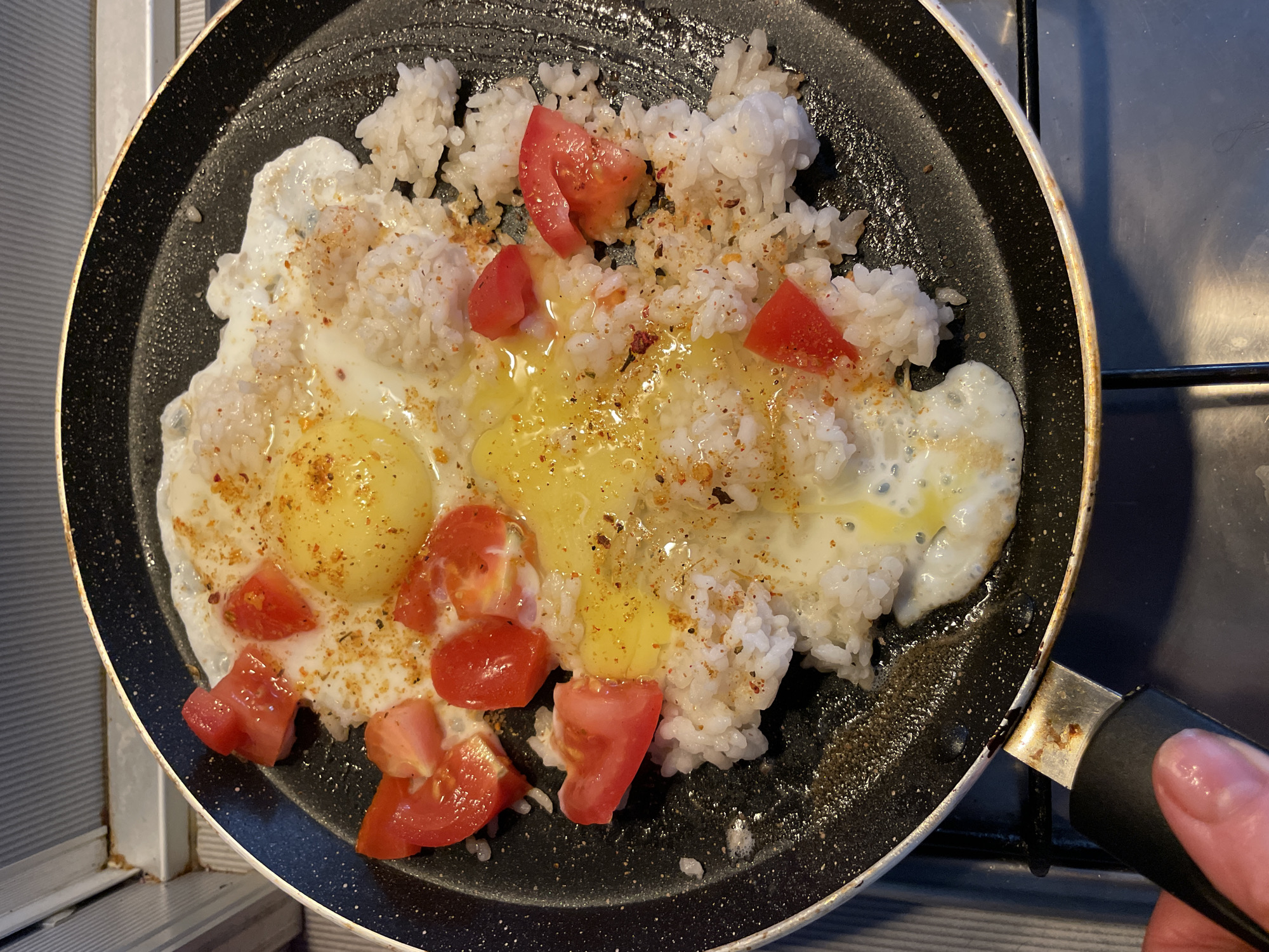 A close-up, overhead shot captures a black frying pan on a stovetop, where a meal is being prepared. The pan contains fluffy white rice interspersed with chunks of bright red tomato and two fried eggs with bright yellow yolks. The eggs are seasoned with a sprinkling of spices that appear to be a mix of red and brown flakes. The rice and eggs are slightly mixed together, suggesting they are being cooked or warmed in unison.
A hand, with a light skin tone and manicured nails, is visible on the right side of the frame, holding the black handle of the frying pan. The hand's position indicates that the pan is being actively managed, possibly stirred or adjusted. The setting appears to be a kitchen, with glimpses of a grey surface behind the pan, likely a stovetop. The lighting suggests it could be daytime, possibly indoors, given the direct overhead view. There is no visible text in the image. The scene is focused entirely on the food preparation, creating a sense of everyday domestic activity.