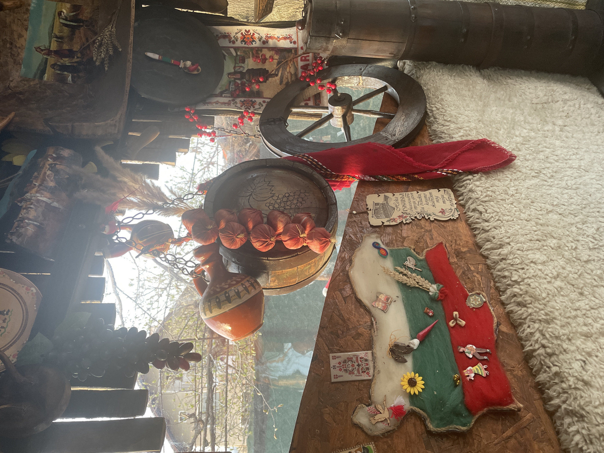 The image shows a still life arrangement in what appears to be a folk-themed interior, possibly in a restaurant or cultural center in Aprilovo, Bulgaria. The composition includes traditional Bulgarian decorative items. Dominating the scene is a handcrafted representation of Bulgaria’s national flag, made from white, green, and red felt or fabric, decorated with small objects like flowers and miniature figures. A red headscarf rests near a wooden wheel and a barrel. Additional items include a ceramic jug, strings of decorative onions, and a framed piece of paper with a building and text. The background shows a glimpse of outdoor foliage through a window.