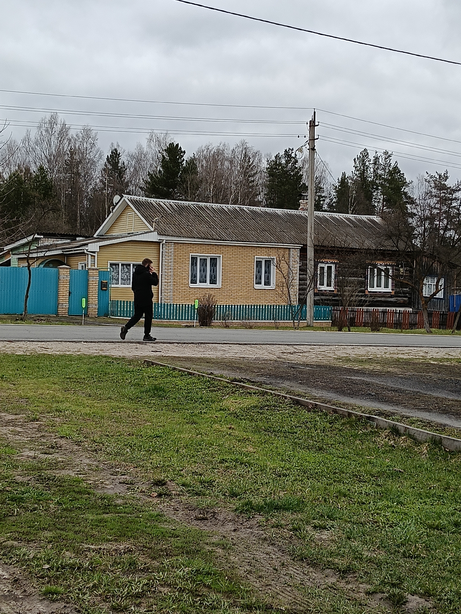 The image shows a man walking along a street in Красный Маяк, Russia. He is wearing a black hooded sweatshirt and dark pants. A wooden house, partially painted yellow brick and partially dark wood, stands behind him. The house has white-framed windows. There is a small fence between the house and the road that has teal-painted sections. A power pole is next to the house. A small blue fence is partially visible to the left. Trees are in the background. The foreground consists of a grassy area with patches of dirt. The sky is overcast.