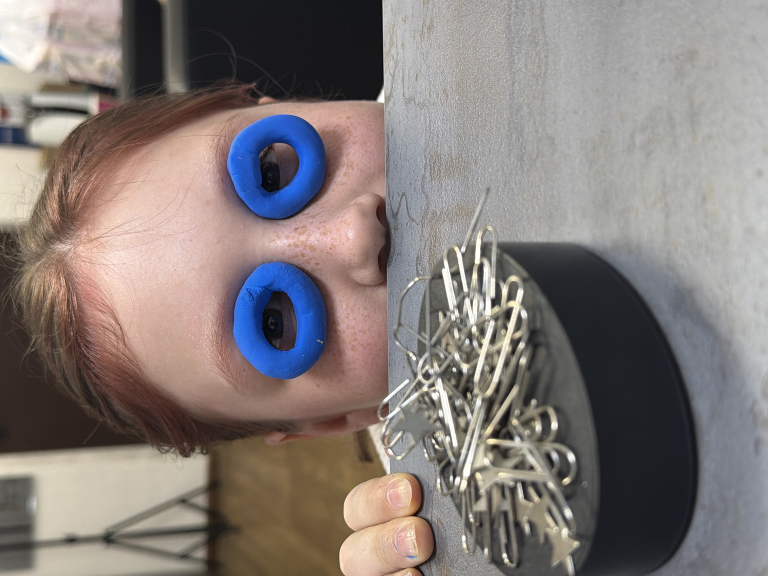 The image is rotated 90 degrees clockwise, presenting a close-up, surreal portrait of a person peeking over an edge, seemingly upside down relative to the image frame. The subject has fair skin with visible freckles on their nose and cheeks, and reddish-brown hair.
Two vibrant blue, toroidal (donut-shaped) objects are placed over the person's eyes, framing them as if they were exaggerated eyelids or goggles. Through the holes of these blue rings, the person's dark eyes are visible and open, looking towards the viewer. The expression is one of curiosity or playful engagement.
To the right of the person's head (in the rotated image), a black cylindrical object, likely a magnetic paperclip holder, is partially visible. It is filled with numerous metallic silver paperclips, including some distinct star-shaped ones. The person's hand, with visible fingernails, is gripping the edge of the surface next to the paperclip holder. The surface appears to be a light-colored, textured wall or desk. The background is mostly blurred, showing indistinct shapes and colors, suggesting an indoor setting.
No visible text is present in the image. The scene takes place in Chaikovsky, Russia.