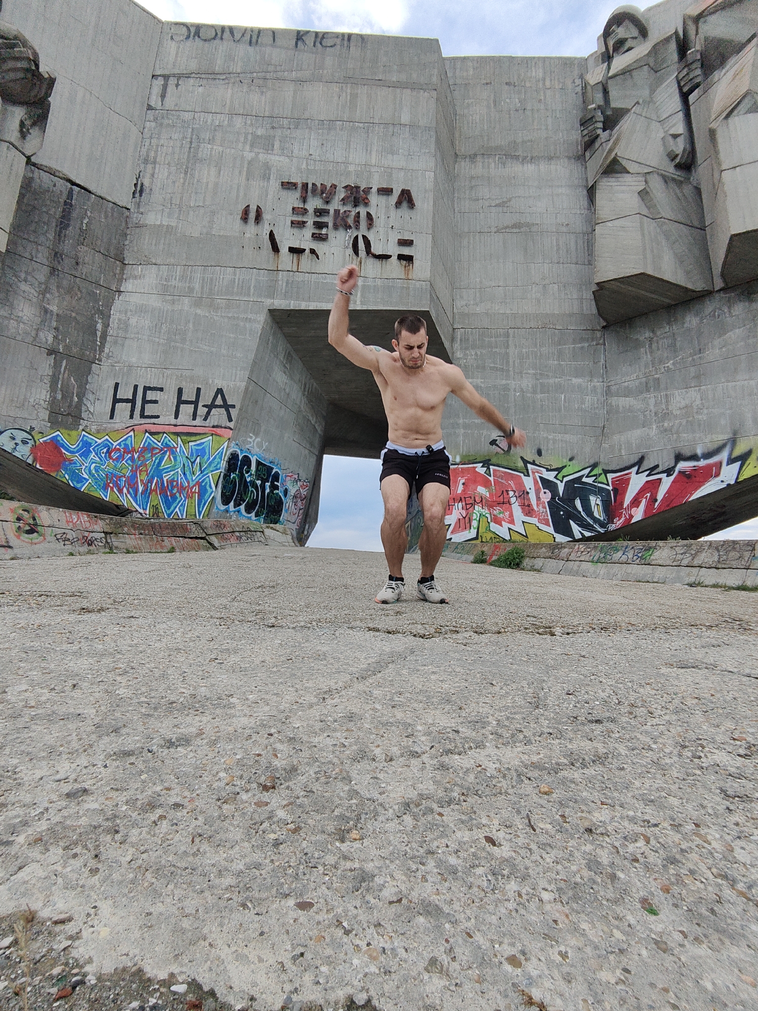 A low-angle medium shot depicts a shirtless man jumping in front of a large concrete structure. The man, who appears to be in his late 20s or early 30s, is wearing black shorts and white sneakers. He is mid-jump, with his right arm raised and his left arm extended to the side.
The background structure is a large, imposing concrete monument covered in graffiti. The words "HE HA" and "DUIVA KICU" are visible on the monument, along with other illegible text and designs. The structure has a large opening in the center, through which the sky is visible.
The ground in front of the monument is also concrete and covered in small stones and debris. The location is the House-Monument of the Bulgarian Communist Party in Buzludzha, Bulgaria.