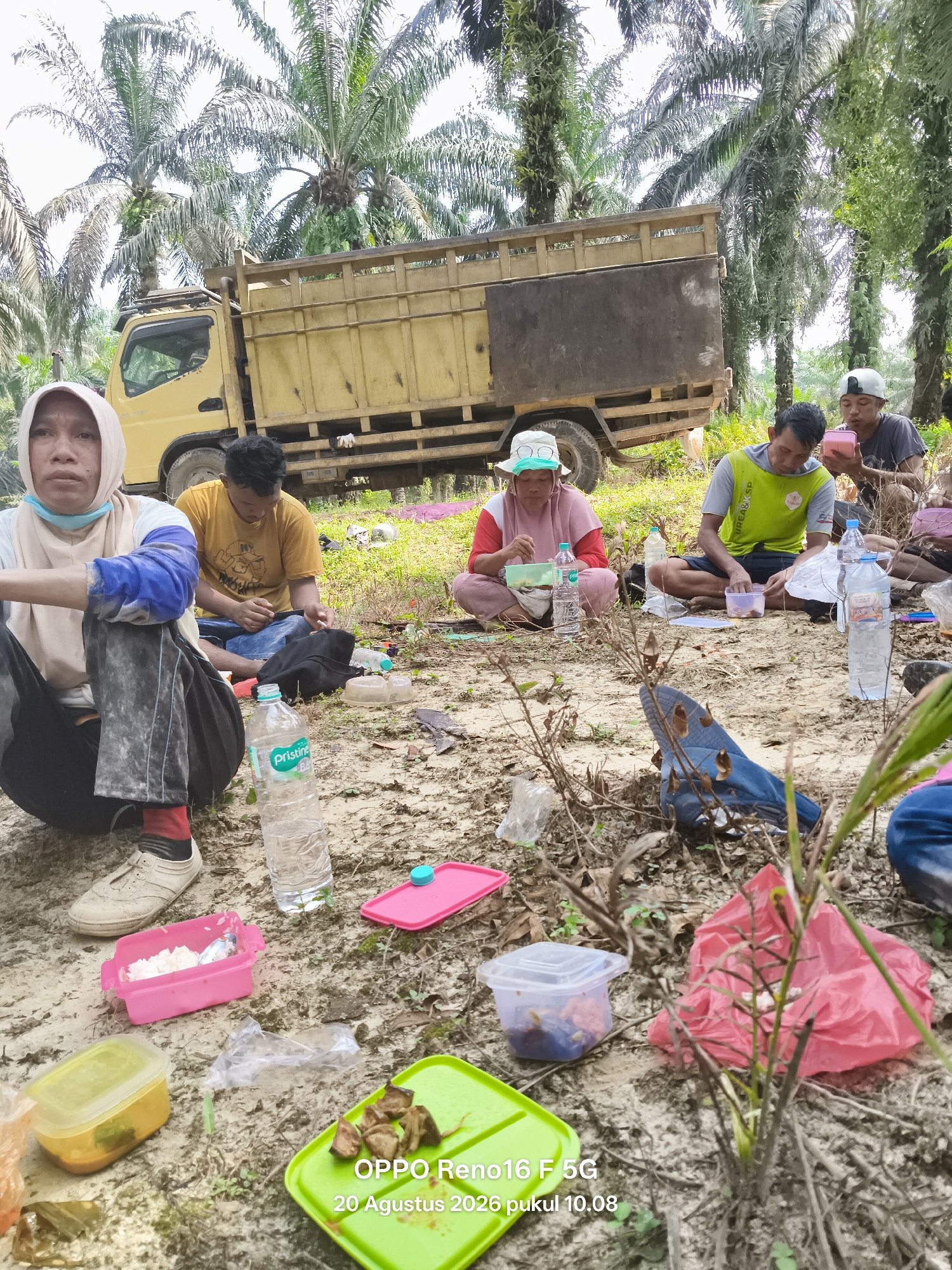 In the city of Penghidupan, Indonesia, a group of individuals are taking a break from work, gathered on the ground amidst a plantation of palm trees. A yellow truck is parked in the background, its bed filled with what appears to be harvested goods. Several people, dressed in casual clothing and some wearing headscarves or caps, are seated around containers of food, engaged in eating or resting. Among the items on the ground are plastic containers of food, water bottles, and a pink plastic bag. The terrain is sandy and covered with dry leaves and vegetation. The overall scene depicts a moment of respite during what seems to be manual labor, likely agricultural work, in an outdoor setting.