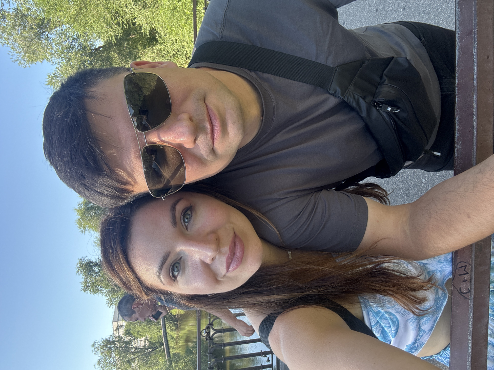 A selfie is taken from a low angle outdoors. The foreground is dominated by the faces of a man and a woman. The man, on the right, is wearing sunglasses and a grey t-shirt with a black fanny pack across his chest. The woman, on the left, has long brown hair and is wearing a blue and white patterned top. Behind them, a body of water with a bridge and trees can be seen. A blurred figure of another person is visible on the bridge. The scene suggests a casual outing in a park or along a river in Kryvyi Rih, Ukraine, on a sunny day.