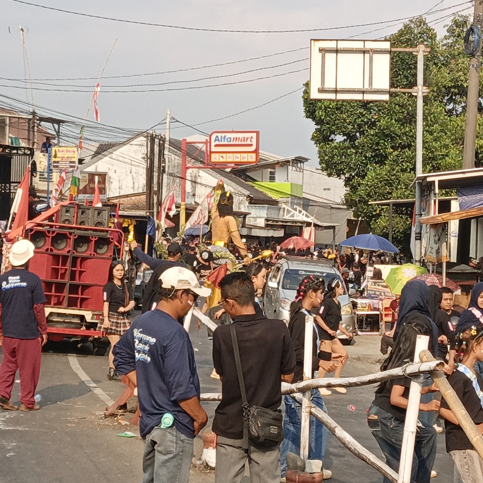 The image depicts an outdoor street scene in Randudongkal, Indonesia, during what appears to be a festive or cultural event. In the foreground, several individuals are visible. A person wearing a dark blue t-shirt with white lettering and a light-colored hat is seen from the back, looking towards a large red speaker system mounted on a vehicle. To their left, another person in a dark blue t-shirt and red pants is also visible.
A prominent red speaker setup, likely for a parade or performance, is a central element. It features multiple large speakers stacked vertically and horizontally. To the right of the speakers, a person in a black t-shirt and a plaid skirt stands, looking towards the left. Further to the right, a woman with dark hair and a black t-shirt is captured mid-motion.
In the middle ground, a crowd of people is gathered. A figure dressed in traditional attire, possibly a performer or effigy, is elevated and prominently displayed, holding a golden object. Several Indonesian flags, red and white, are visible throughout the scene, attached to poles or held by individuals, suggesting a national celebration or event. Umbrellas, some blue and some green, are open, indicating potential shade from the sun or protection from light rain.
The background reveals a street lined with buildings, characteristic of a town or city environment. A red sign with white lettering, appearing to be an "Alfamart" logo, is visible above the crowd, indicating a commercial presence. Overhead power lines crisscross the sky, a common sight in urban Indonesian landscapes. The ground is paved and appears somewhat littered. The overall atmosphere suggests a lively public gathering.