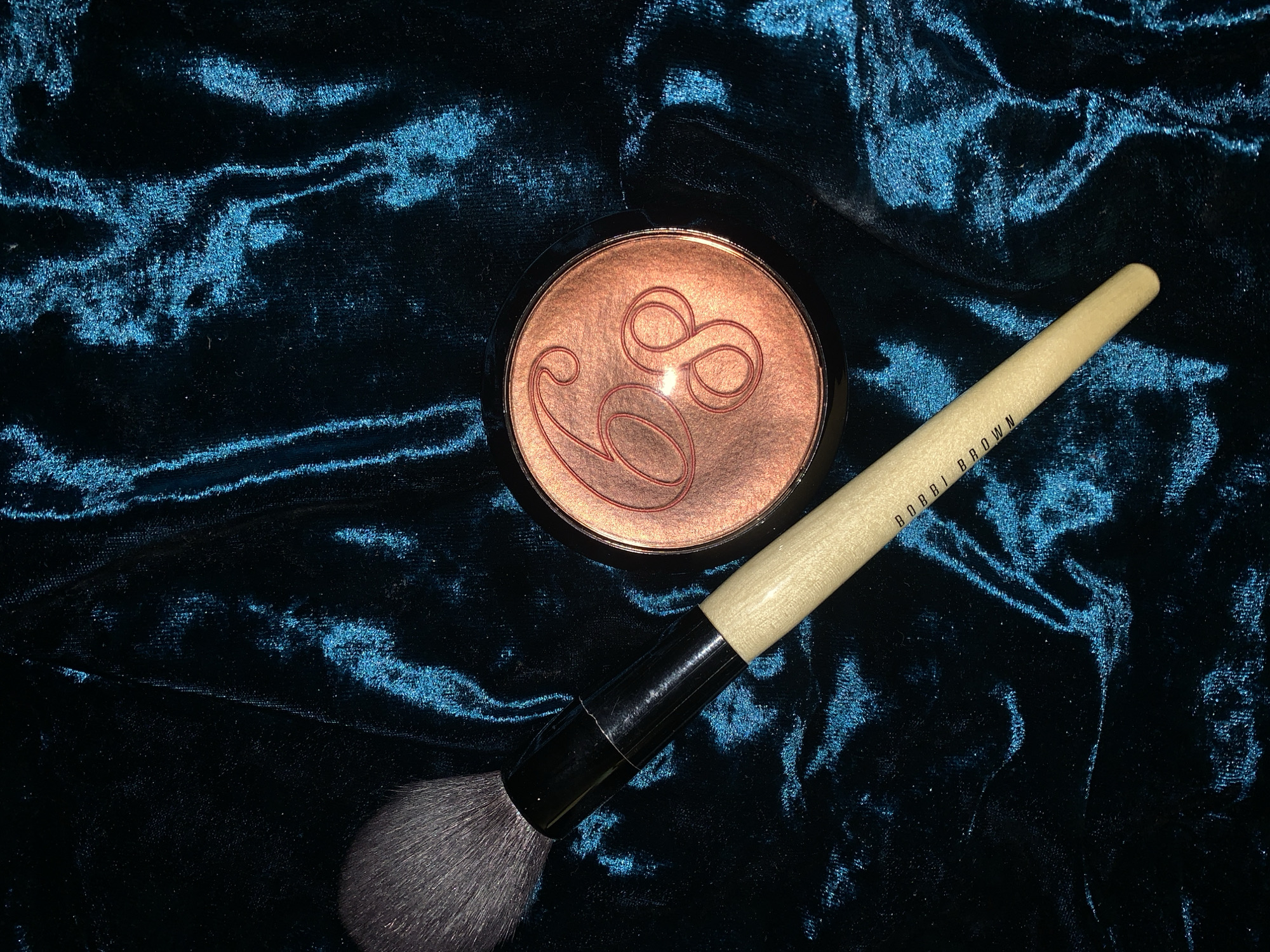 A makeup brush and a compact of bronzer are laid out on a dark, teal-colored velvet fabric. The brush has a light-colored handle with "BOBBI BROWN" printed in black text. The bronzer compact is round with a copper-toned powder inside, embossed with a stylized "68". The velvet fabric has a high sheen and appears to be draped, creating folds and highlights.