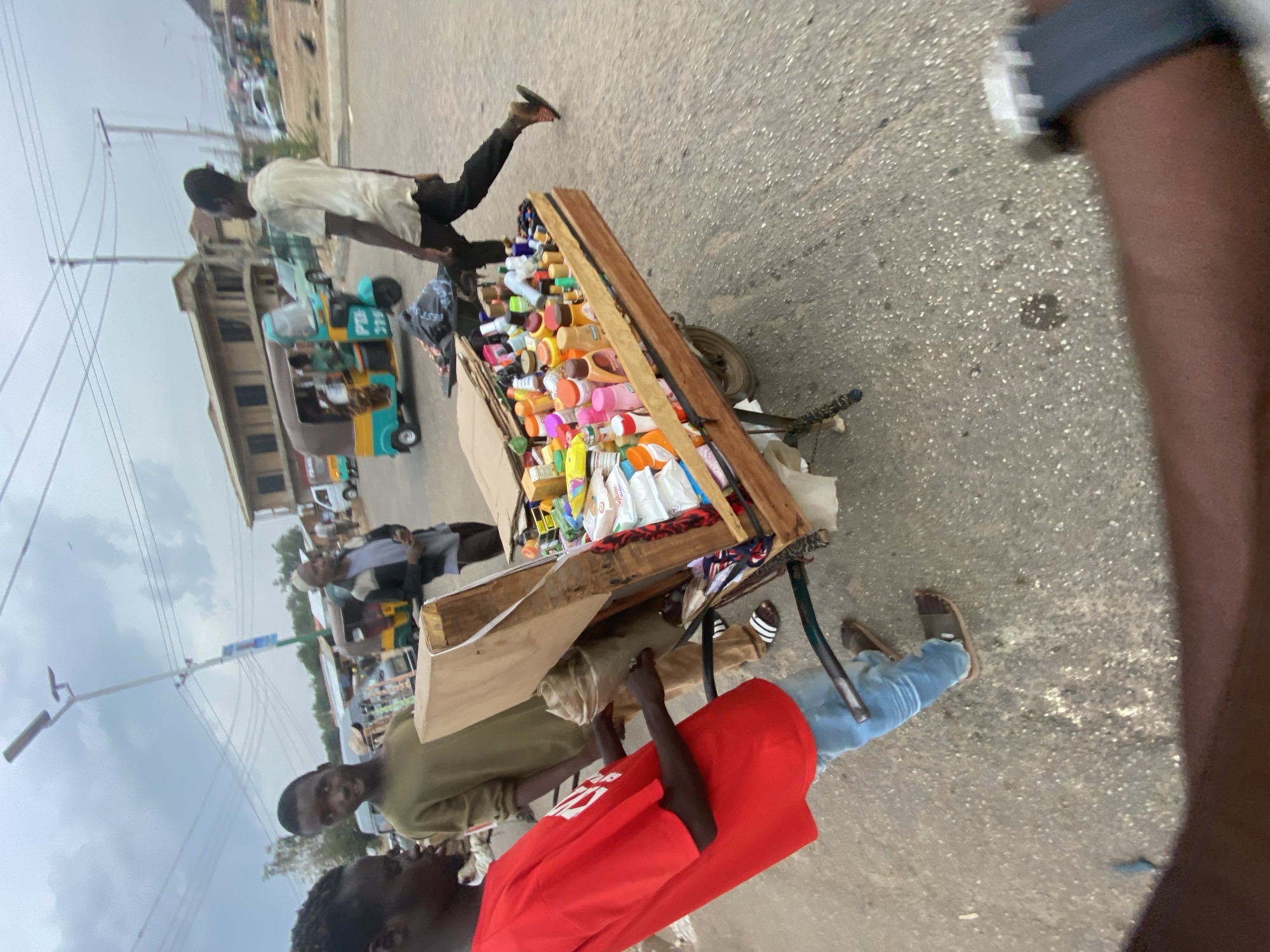 This media file captures a street scene in Bukuru, Nigeria, featuring several individuals and a vendor's cart under an overcast sky. The image is taken at an oblique angle, looking down a paved street.
In the foreground, a wooden pushcart is laden with numerous brightly colored bottles and packages, likely cosmetics or small household items, suggesting a street vendor operation. Two individuals are prominently positioned near the cart. One person wears a red t-shirt with white text (illegible) and blue jeans, standing with their back mostly to the camera. Another person, wearing an olive green shirt, stands opposite, looking towards the camera with a subtle expression.
Further down the street, a person in a light-colored shirt and dark trousers walks away from the camera, past the cart. In the mid-ground, a multi-colored auto-rickshaw, commonly known as a Keke NAPEP, is visible with partial text like "P.T.S." on its side. Other people are discernible further in the background, walking or standing by buildings.
The setting includes power lines and utility poles against a cloudy sky, indicating daytime. Buildings line the street in the distance. The overall scene depicts a bustling, everyday street environment characteristic of an urban or semi-urban area in Nigeria, with active street vending and transportation. Specific emotions of the individuals are not clearly discernable due to distance and image quality.