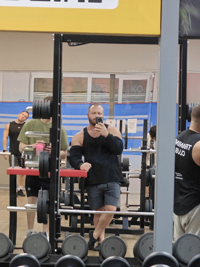 The image depicts an indoor fitness center located in Chaikovsky, Russia, primarily viewed through a large mirror. A man with a bald head and a beard, wearing a black sleeveless shirt, black arm sleeves, grey shorts, and dark shoes, is taking a selfie with a dark smartphone held to his face. He is leaning his left arm on a red-topped rack support, with his legs crossed at the ankles.
Reflected in the mirror, to the man's left, another male is visible wearing a black tank top and dark shorts, looking upwards and to the left. Partially obscured behind a black squat rack structure, another male wearing an olive green t-shirt and dark shorts holds a pink-capped bottle and a phone. To the far right, a partial reflection of a fourth individual shows the back of a black tank top with "TRAINING CLUB" printed in white.
The gym floor is light-colored, and the background features large windows revealing an exterior view, a blue and white striped wall or banner, and various wall signs, one with the partial word "KO". Several black and silver weighted barbells are racked on black metal structures. In the immediate foreground, a rack of dumbbells is visible, some marked with "8" and the brand "TECHNOGYM". An overhead yellow structure is also present.