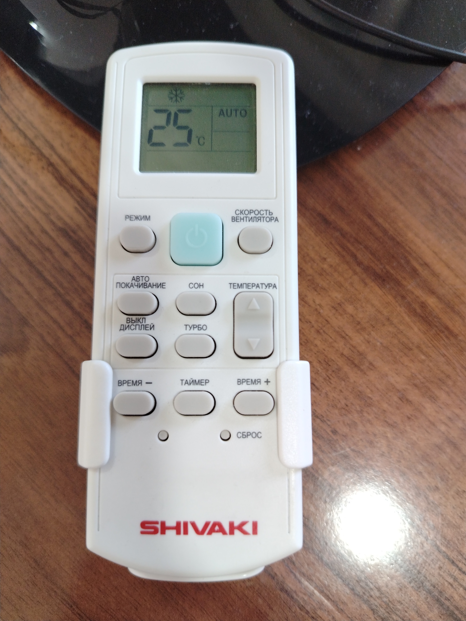 An overhead, eye-level shot shows a white remote control for an air conditioner on a wooden desk. The remote is oriented vertically, with its top towards the top of the image. The display screen at the top of the remote shows "25" with a snowflake icon to its left and "AUTO" to its right. Below the display are various buttons, including a power button in light blue, and several gray buttons labeled in Russian. The brand name "SHIVAKI" is printed in red letters at the bottom of the remote. The wooden desk surface appears to be a light brown color with a visible wood grain pattern. A black object, possibly the base of a monitor or speaker, is visible in the upper portion of the image, partially obscuring the background. The lighting suggests an indoor environment. The image does not depict any people or any specific landmarks or activities that would associate it with the city of Nurata, Uzbekistan.
