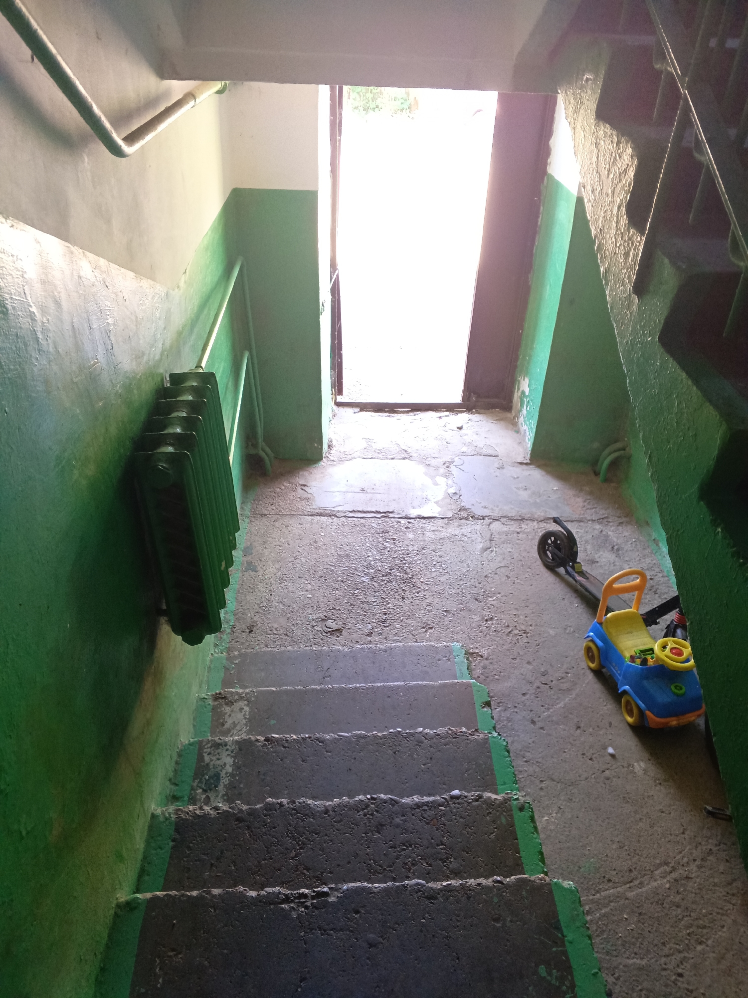 The image shows a poorly maintained stairwell in a building in what is possibly Tolbazy, Russia. The walls are painted in a two-tone color scheme of white above and green below. The paint is peeling and worn. A green radiator is mounted on the wall next to the stairs. The concrete stairs are worn and the edges are painted green. At the bottom of the stairs, near the doorway leading outside, are a child's blue and yellow ride-on toy car and a black scooter. The doorway is open, allowing bright light to illuminate the landing. A metal pipe runs along the ceiling.