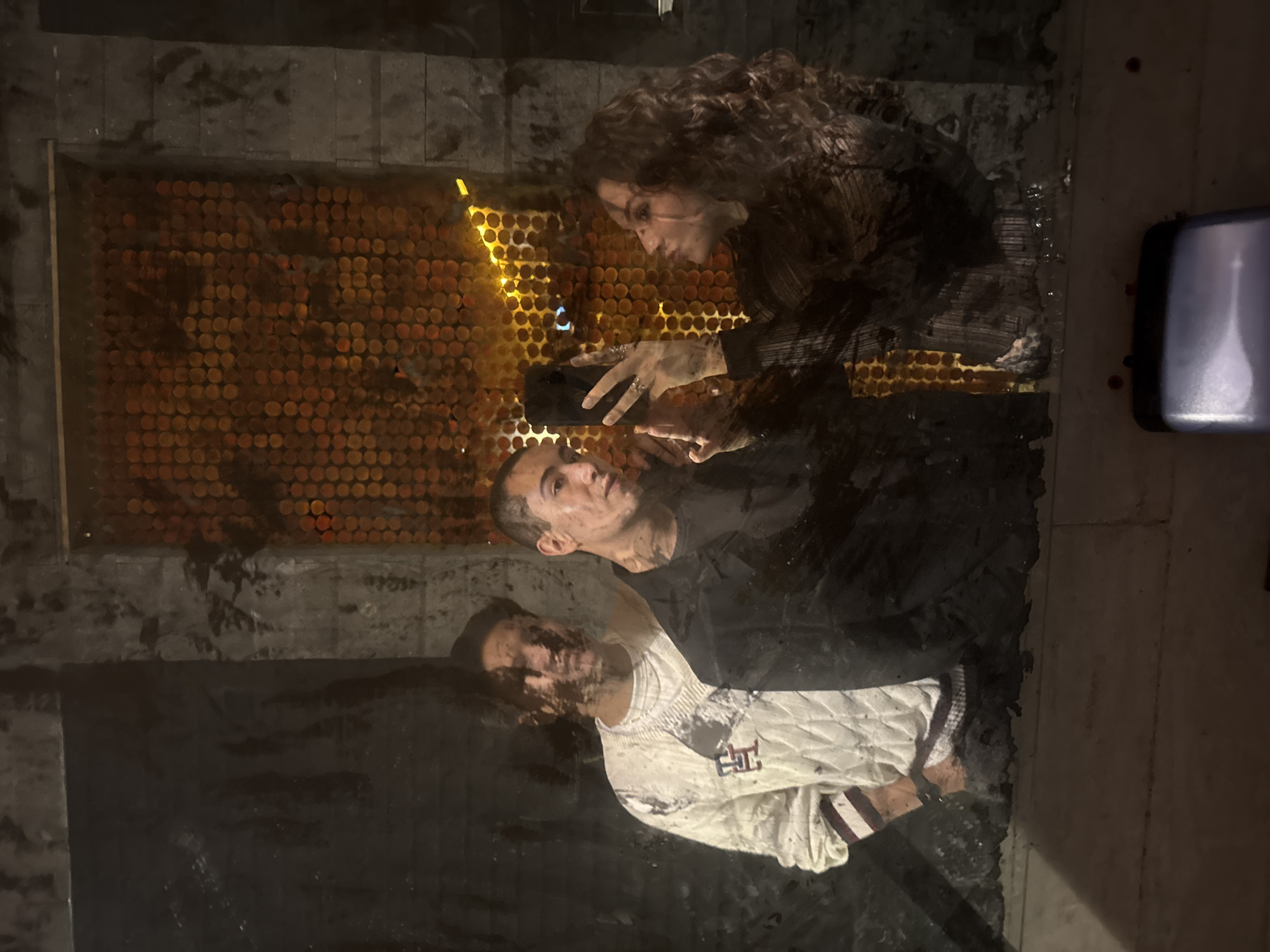 A photograph taken in San Andrés Cholula, Mexico, presents a complex scene, likely a reflection captured on a wet or polished surface. Three individuals are discernable. A woman with curly hair is visible, appearing to hold a mobile phone. Below her, a man is seen, possibly looking up at the woman. A third person is at the bottom of the reflection, wearing a white and red sweater. Behind the reflected figures, there is an illuminated wall containing round fixtures. The surrounding area appears to be urban, with dark and possibly wet surfaces, and a piece of mechanical equipment on the right of the image.