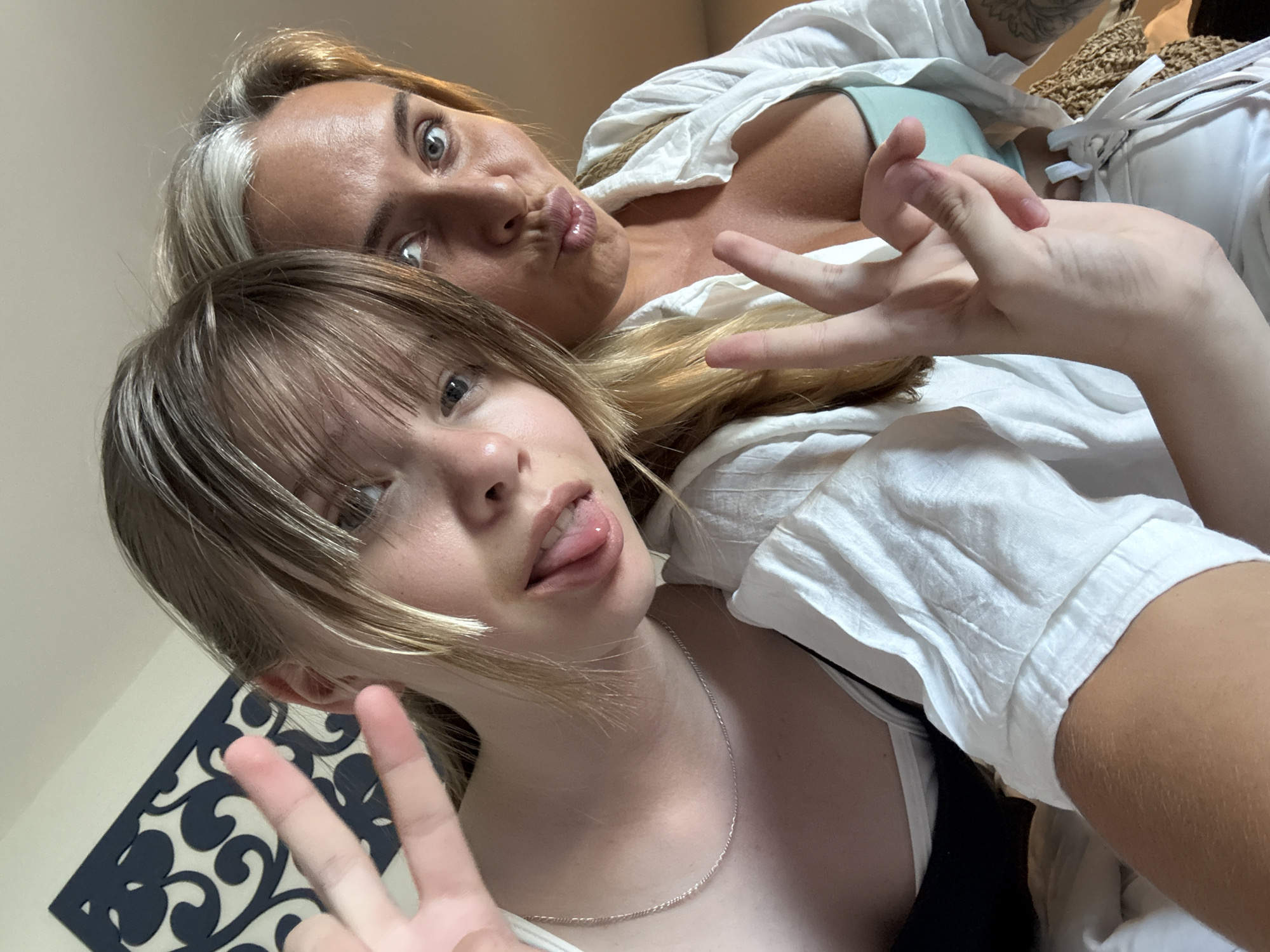 Two people, likely a mother and daughter, are taking a selfie. The woman in the foreground has blonde hair with some grey streaks and is sticking her tongue out. She is wearing a white shirt and a turquoise bra. The person behind her has blonde hair and is wearing a white shirt. They are both looking at the camera. The background is out of focus, but appears to be an indoor setting. There is no visible text in the image. The image appears to be taken during the day.