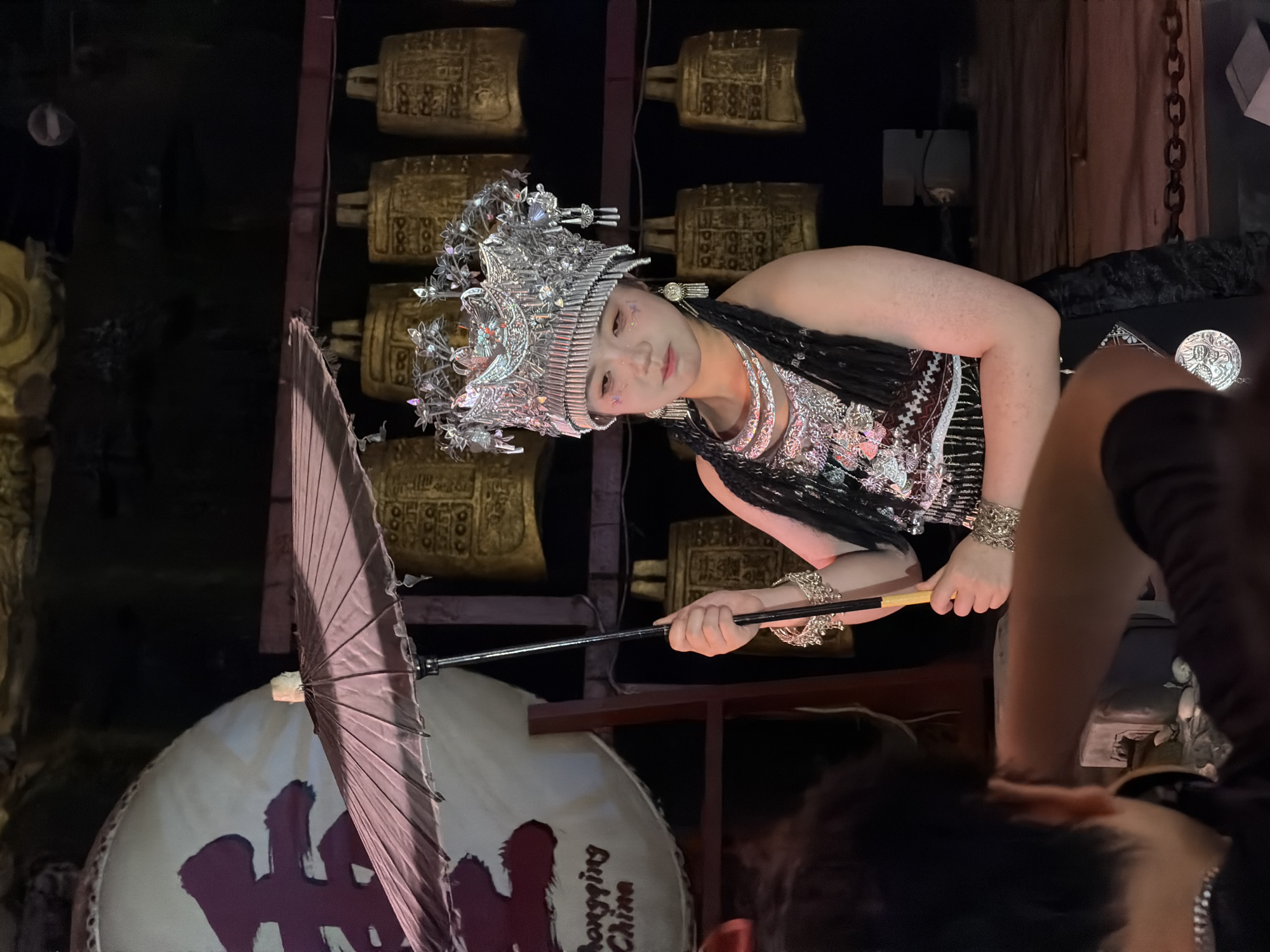 A woman in traditional attire stands holding a closed umbrella, looking downwards. She wears an elaborate headdress adorned with silver ornaments and beaded tassels, a layered necklace, and bracelets. Her outfit includes a patterned vest and a black skirt with a velvet trim. She has glittery makeup on her face. In the background, a large white drum with red Chinese characters is visible, along with a series of golden bells. The scene appears to be a performance or cultural display, likely in the Yuzhong District of Chongqing, China, given the text on the drum. A person's back and shoulder are visible in the foreground, obscuring part of the woman and her lower body. The lighting is dim, highlighting the subjects and the golden bells.