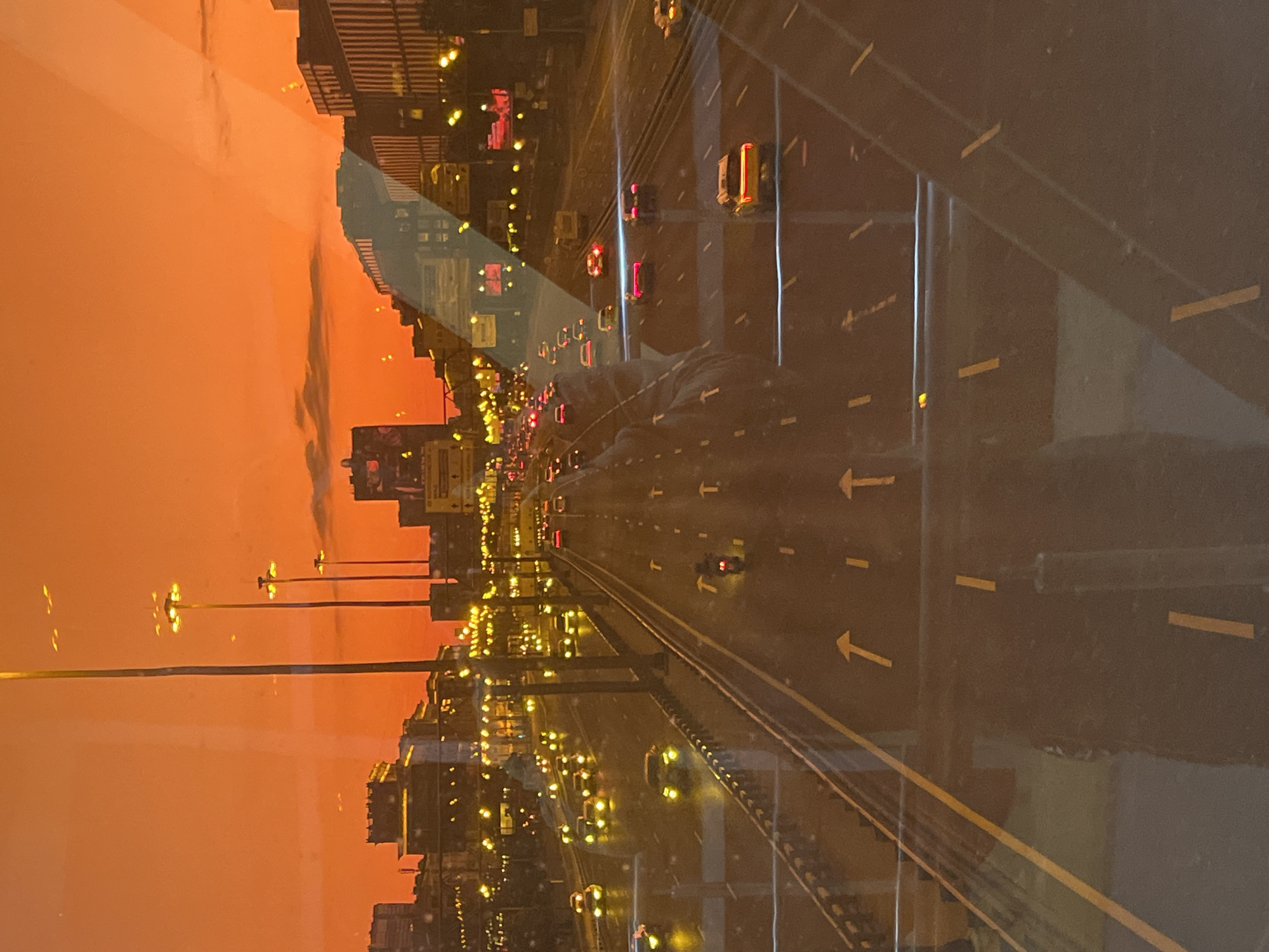 A nighttime view of a multi-lane highway in Khimki, Russia, is captured, likely from an elevated perspective. The sky is a vibrant orange, indicative of either sunset or sunrise. Streetlights line the highway, casting a yellow glow on the asphalt and the vehicles. Several cars and a motorcycle are visible on the road, their headlights and taillights illuminating the scene. Buildings with illuminated windows are present in the background, suggesting an urban environment. The image appears to be a reflection, with a faint human silhouette visible in the foreground.