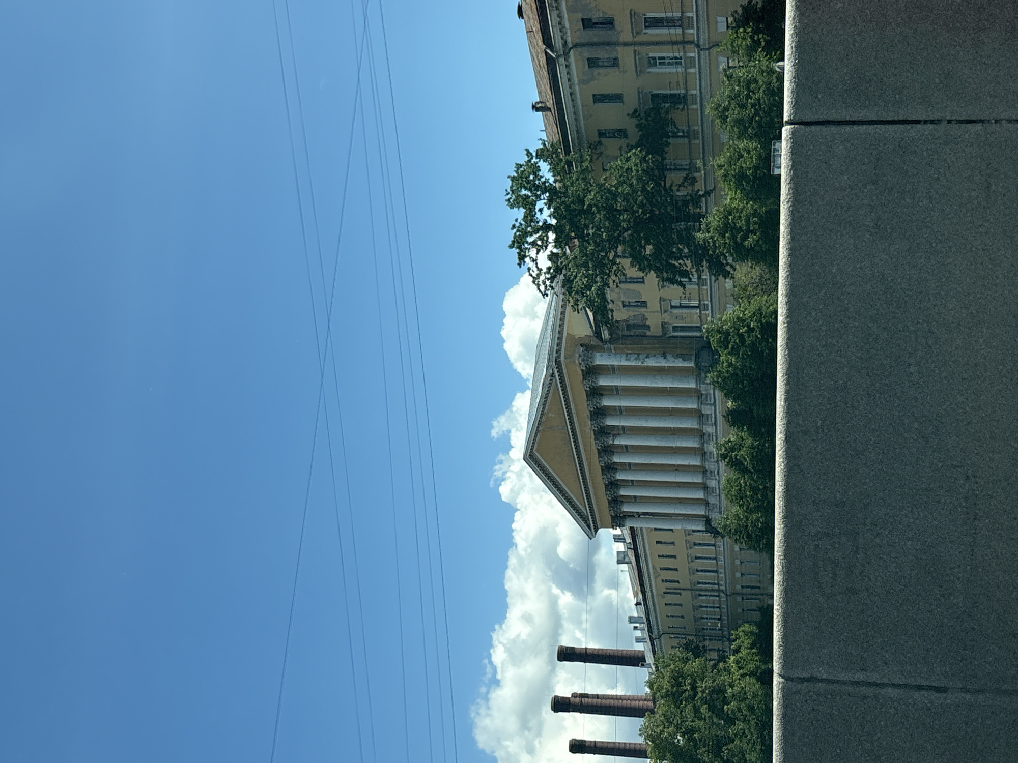 The image shows the building of the former Russian State Optical Institute, located on the banks of the Fontanka River in Saint Petersburg, Russia. The building is a long, yellow structure with classical architectural features, including a portico with Corinthian columns and a triangular pediment. To the left of the main building, three tall brick chimneys rise against the blue sky, indicating the presence of former industrial activity. Dense green trees partially obscure the facade of the building, suggesting it is surrounded by greenery. The sky is bright blue with scattered white clouds, and power lines are visible across the upper portion of the frame. In the foreground, a grey concrete barrier runs horizontally across the image, obscuring the lower part of the scene. There are no people visible in the image. The overall impression is of a historic, somewhat weathered urban landscape bathed in sunlight.