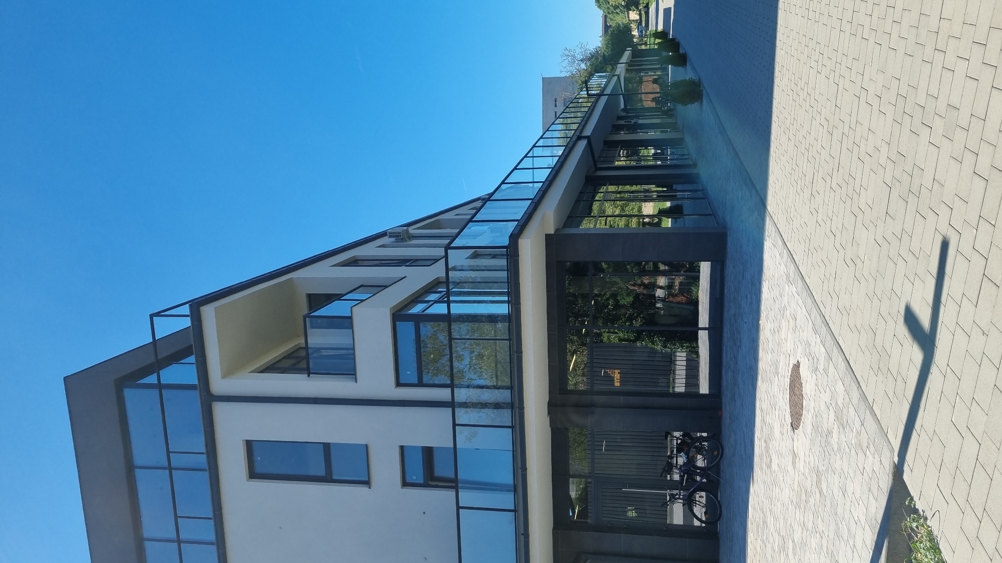 The image depicts a modern, multi-story building with a light-colored facade, dark gray trim, and large glass windows and balconies with glass railings. The building is situated on a paved walkway made of rectangular gray bricks. To the left of the building, two bicycles are parked in front of a dark fence. The building appears to be a commercial or residential property. The sky is clear and blue, indicating a sunny day. In the background, a street lined with trees and other buildings can be seen, suggesting an urban environment. The overall scene is clean and well-maintained. Based on the information available, this location is in the city of Berehove, Ukraine.