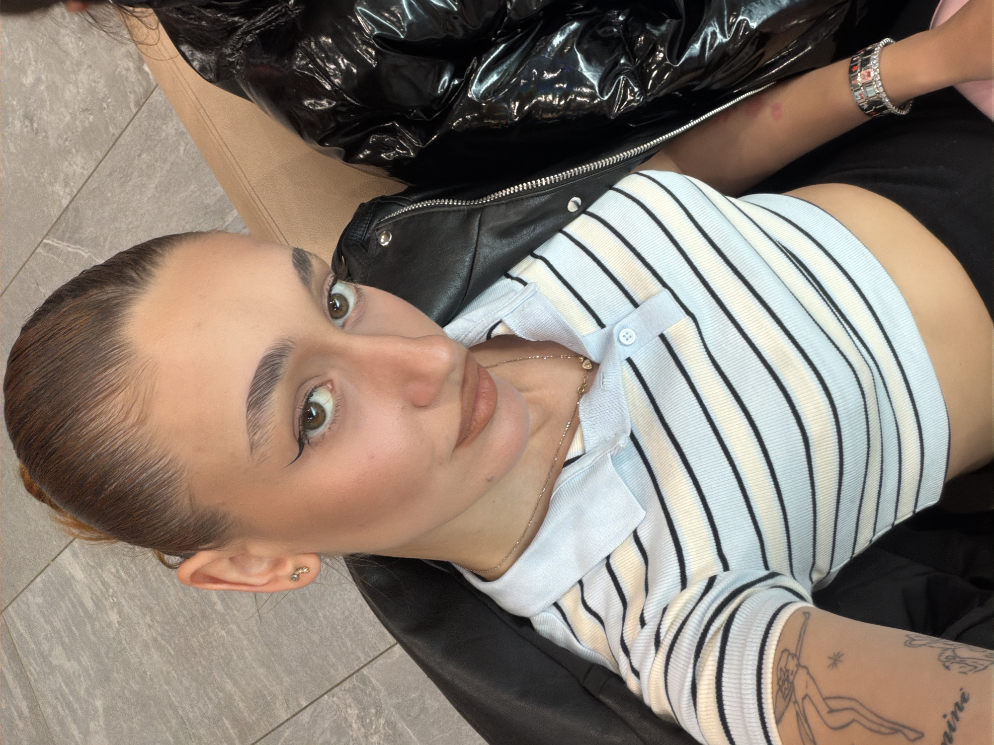 A woman is lying back, looking up at the camera. She has brown hair pulled back, fair skin, and light makeup. She's wearing a striped shirt with a collar and a delicate necklace. She has a leather jacket nearby and a bracelet on one wrist. Part of a shiny, possibly vinyl, puffer jacket is also visible. The background consists of a grey tiled floor and the corner of a light-colored piece of furniture. There is not enough information in the image to determine the location in Sofia, Bulgaria.
