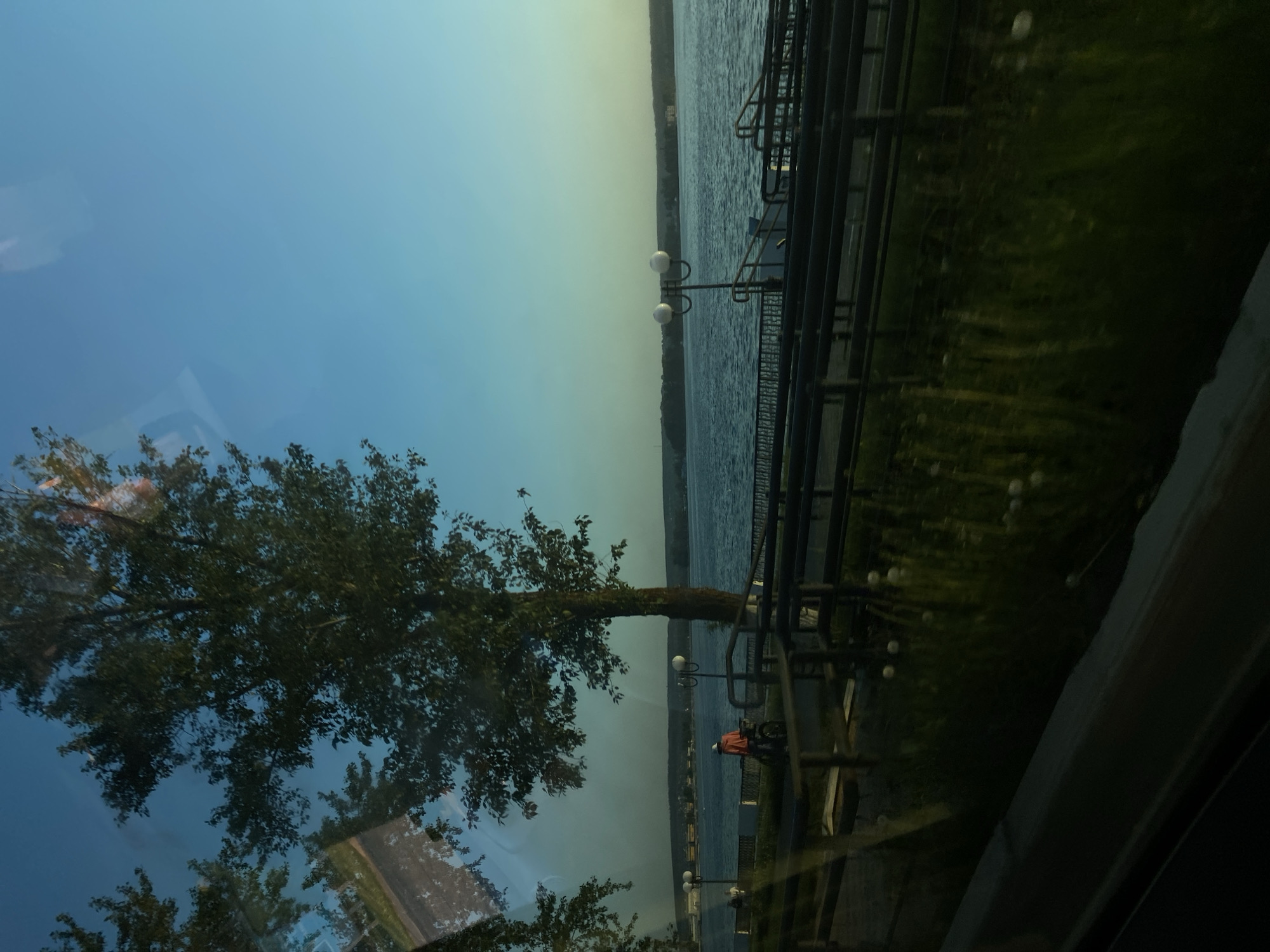 The image captures a scene at the Votkinsk reservoir in Votkinsk, Russia, likely viewed through a window. The reservoir dominates the background, extending to the horizon under a gradient sky shifting from blue to a muted yellow. A tree is visible in the foreground, its branches partially reflecting onto the glass surface. A black metal fence and streetlight line the edge of the water. A person with an orange shirt can be seen on the embankment near the water, in front of the tree.