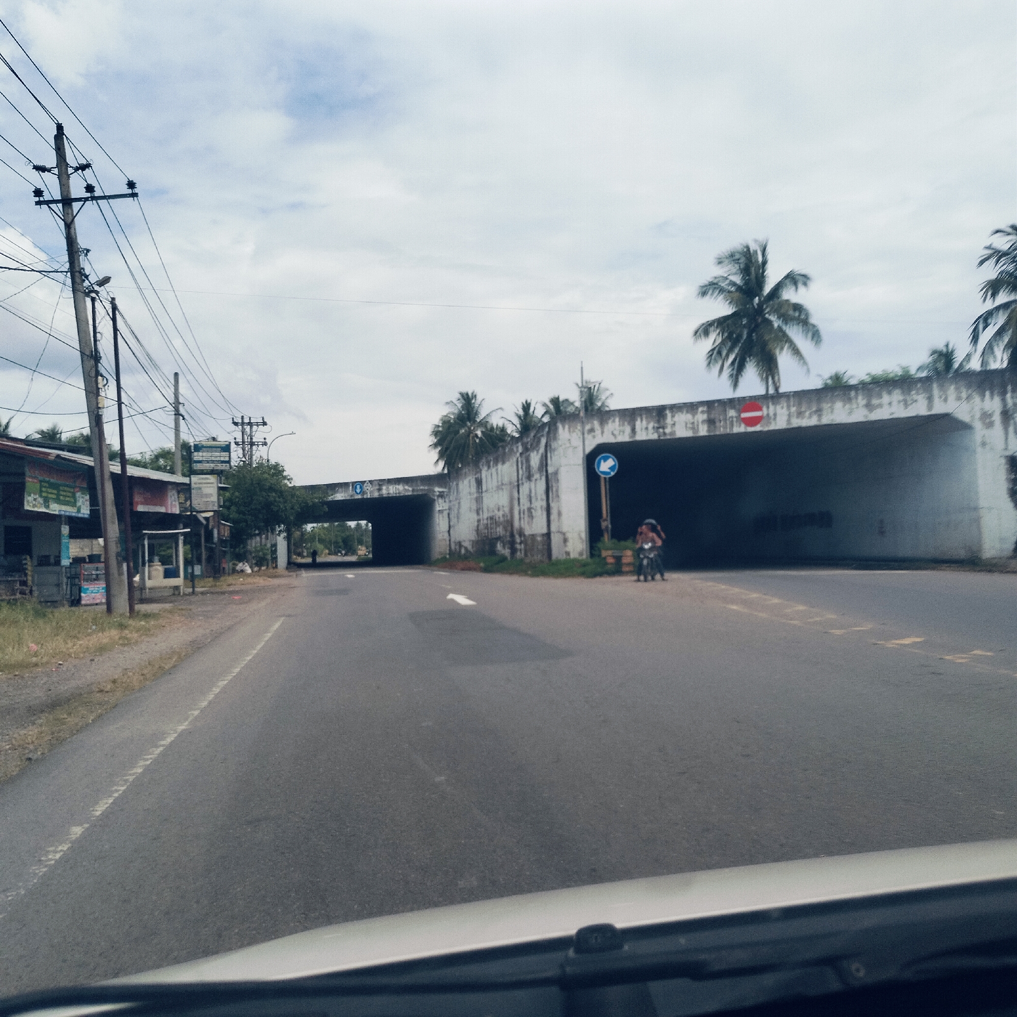 The image depicts a road scene in Cottunong, Indonesia, viewed from inside a vehicle, looking towards a large concrete underpass or tunnel system. The sky is overcast, suggesting a cloudy daytime.
In the foreground, an asphalt road with a white directional arrow painted on it leads towards the underpass. On the left side of the road, multiple utility poles with tangled overhead wires stand beside a row of simple roadside buildings or stalls. While text on these signs is largely illegible due to distance, some appear to feature Indonesian script.
The prominent feature is a wide, weathered concrete underpass structure spanning the width of the road, featuring two large, dark openings. Palm trees grow on top of and behind this structure. Above the left underpass opening, a blue circular sign with a white arrow pointing right is faintly visible. Above the right underpass entrance, a red circular "do not enter" sign is clearly seen. Near this right entrance, a person on a motorcycle is visible, next to a blue circular sign with a white left-pointing arrow. The overall scene suggests a functional, bustling road environment common in semi-urban Indonesian areas.