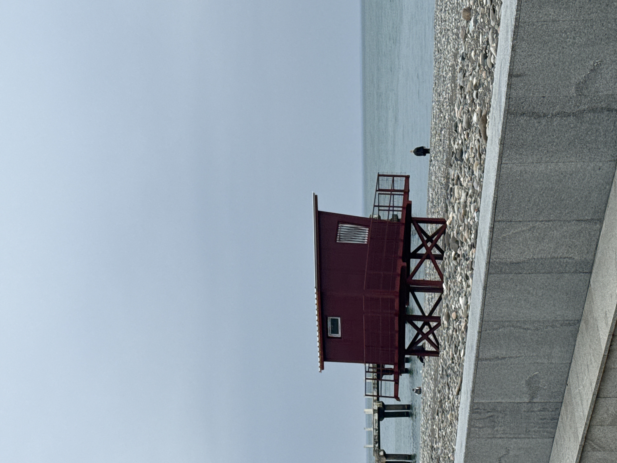 The image depicts a maroon wooden structure on stilts, positioned along the coastline of Batumi, Georgia. The structure, possibly a lifeguard station or a fishing shack, features a small enclosed space with a barred window and a smaller, square window below. It is situated on a pebble beach beside the Black Sea. A concrete wall or embankment runs along the edge of the beach. There is a solitary dark bird visible on the beach, near the structure. The sky is overcast and pale, suggesting a hazy day.