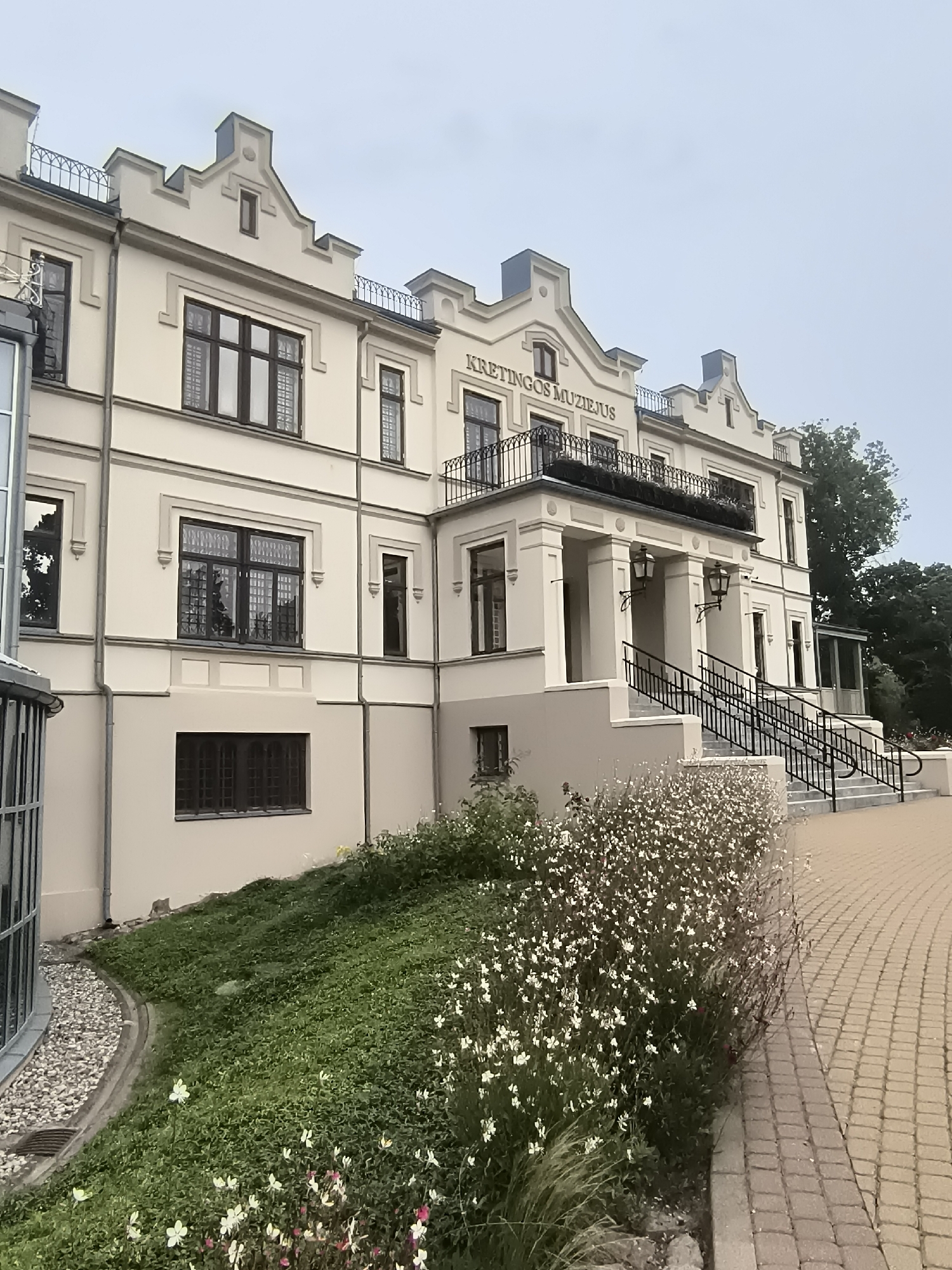 The image shows the Kretingos Muziejus, a museum located in Kretinga, Lithuania. The museum is a large, cream-colored building with a distinctive architectural style, featuring gabled roofs, ornate window frames, and decorative railings. A wide, paved walkway leads to the entrance, which is accessed by a grand set of stairs flanked by planters and lampposts. The foreground is filled with lush green grass and a dense bed of white and pink flowers. The sky is overcast. The image is a medium shot, capturing the building's facade and its surrounding landscape.