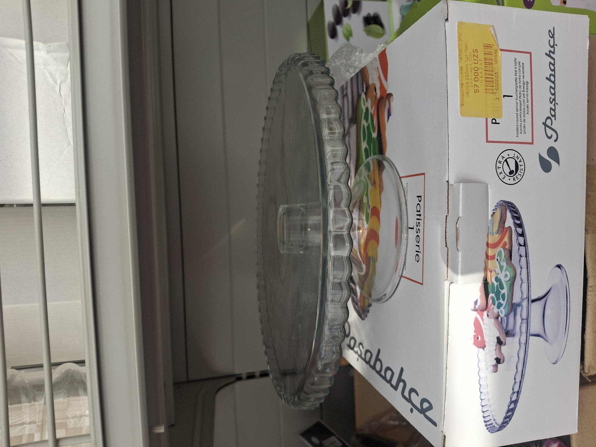 A glass cake stand with a fluted rim and a flared base is displayed on top of a cardboard box. The box has printed images of cookies on it and text in several languages, including Russian and English. A yellow price tag is attached to the box. In the background, there are shelves with items that appear to be in plastic packaging. The scene is lit with artificial light. The overall impression is that of a retail environment, likely a store or warehouse. The presence of Russian text and Cyrillic script on the price tag suggests a region where Russian is spoken or understood, such as Uzbekistan, where Cyrillic script is still in common use. The location of Tashkent, Uzbekistan, is not explicitly evident from the image itself but is provided as a context.