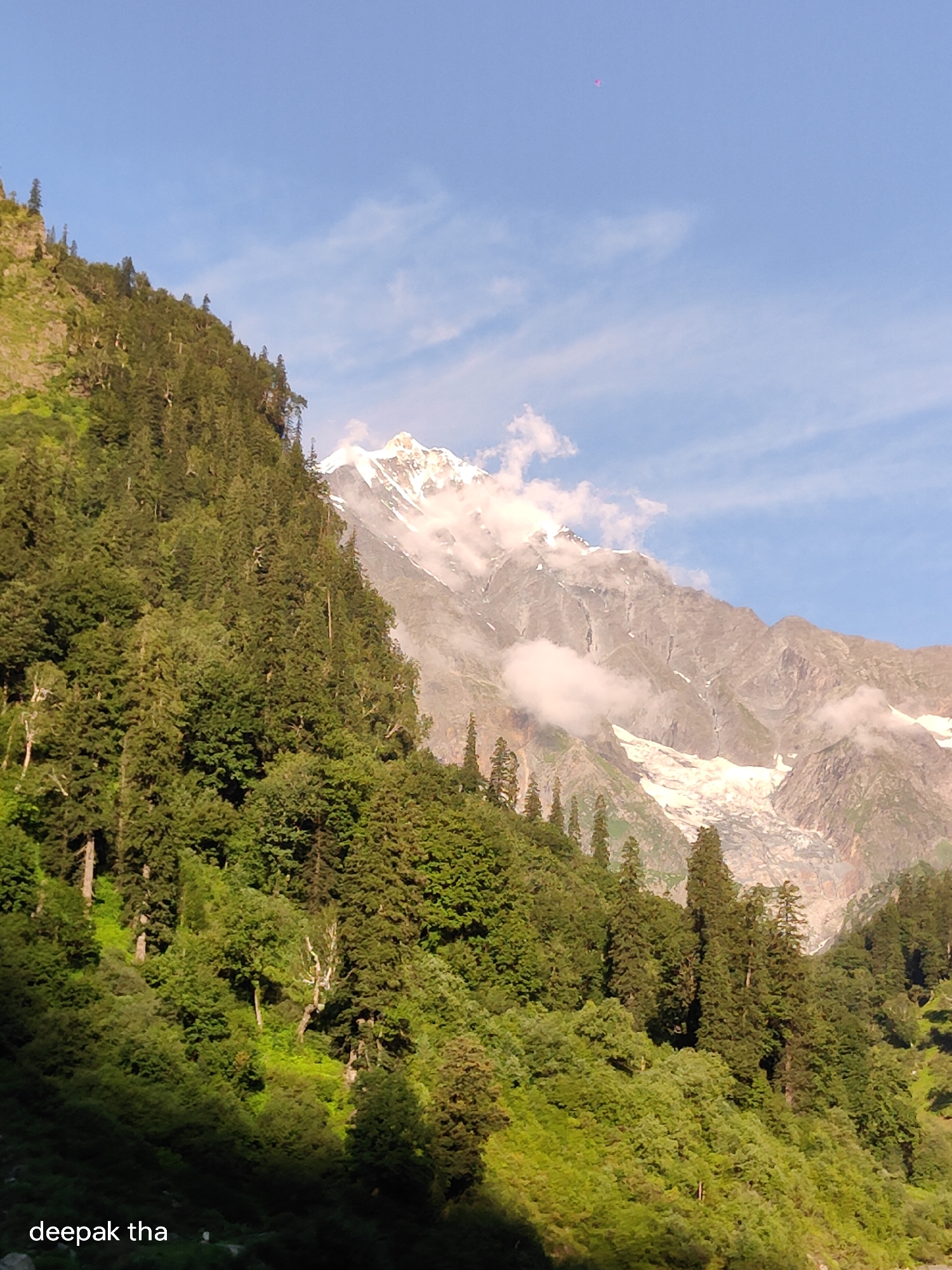 A full shot captures a sun-drenched mountain range partially obscured by mist and snow. Dense green trees cover the lower slopes of the mountains. The sky is a clear blue with a few wispy clouds. The text "deepak tha" is visible in the lower left corner of the image, possibly indicating the photographer or a watermark. The image is likely taken in the region of Burwa, India, given the style of the landscape and vegetation.