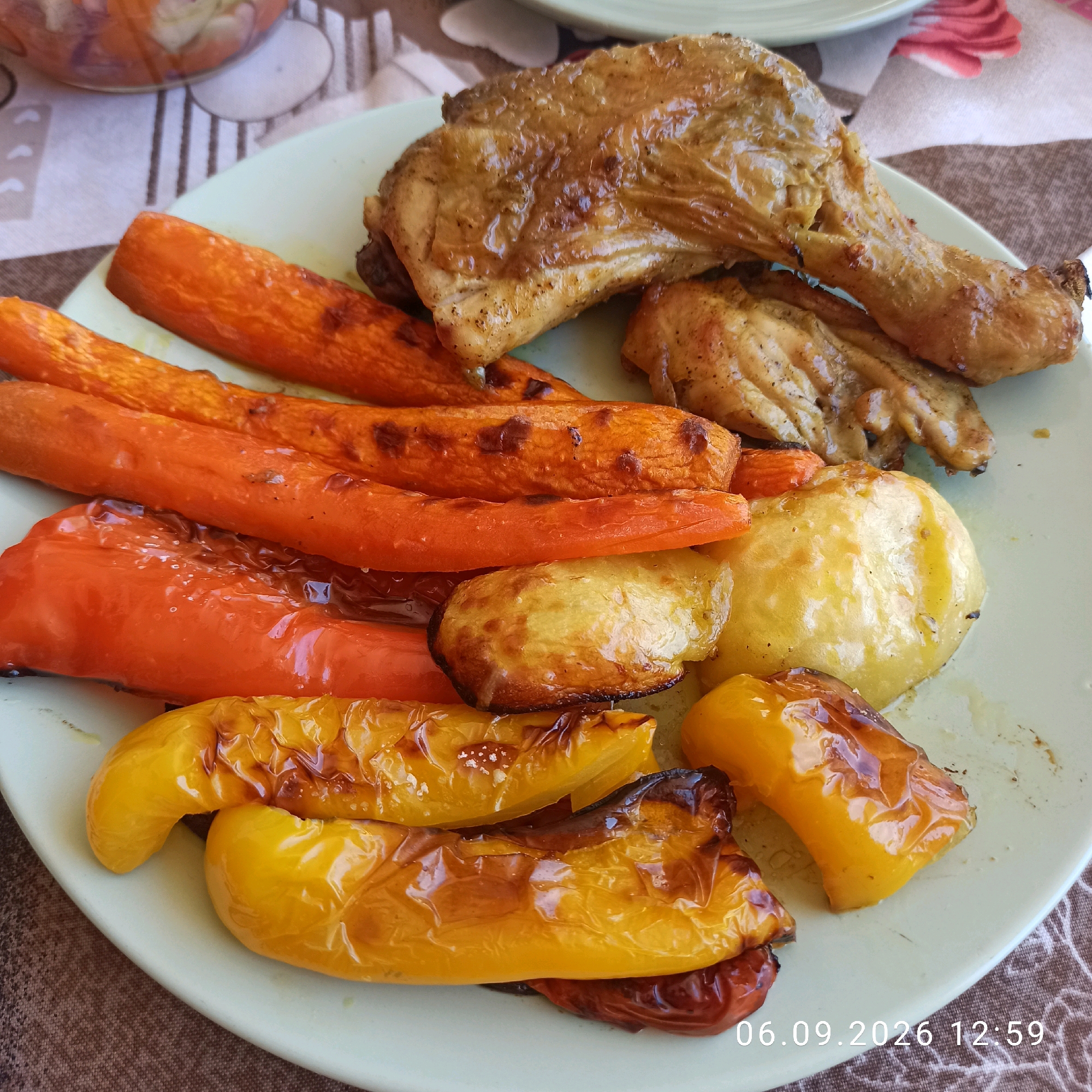 A close-up shot showcases a light green plate filled with roasted chicken pieces, roasted carrots, roasted bell peppers, and roasted potatoes. The food appears to be glazed and glistening, suggesting it has been recently cooked and seasoned. The background reveals a brown and white patterned tablecloth, with a blurred glass bowl containing salad visible in the upper left corner. A timestamp in the lower right corner reads "06.09.2026 12:59." The image is set on a table. There is no information in the image to indicate the location in the city of Bitonto, Italy.